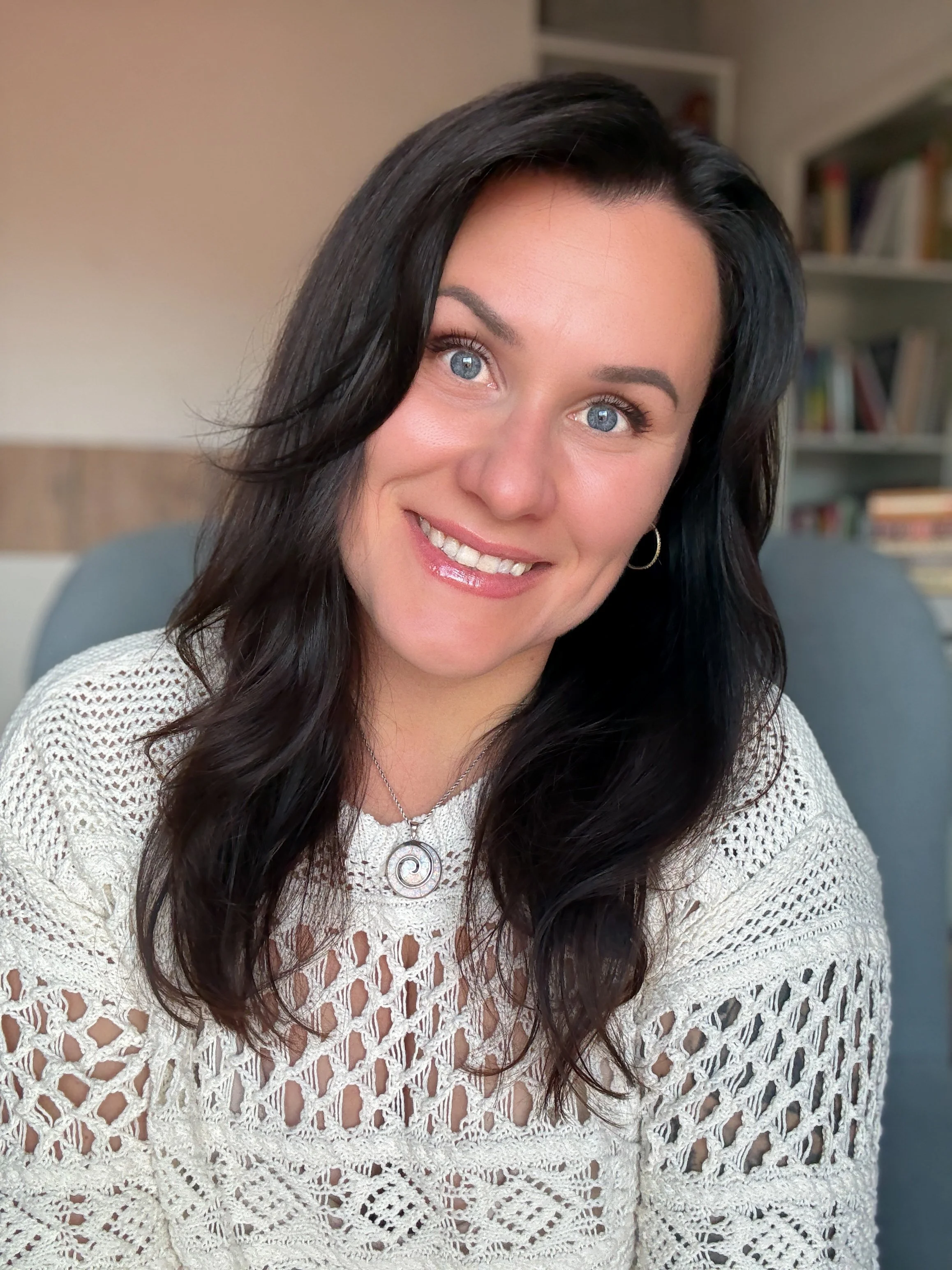 Close-up of a woman with long dark hair, blue eyes, wearing a white crochet top and a circular pendant necklace, smiling indoors in front of a bookshelf.