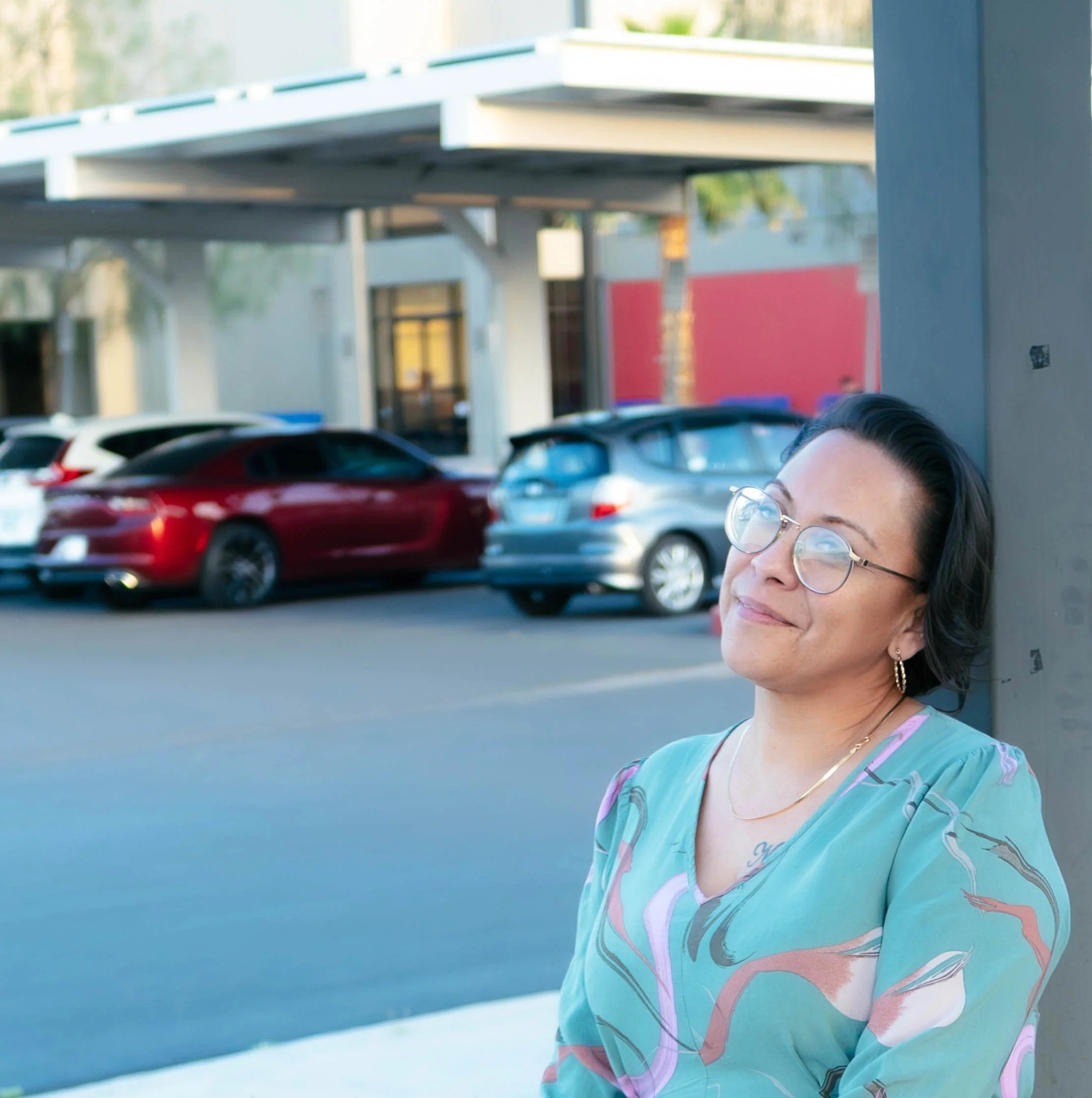 Woman with glasses and earrings leaning against a wall outside, with parked cars and a building in the background.