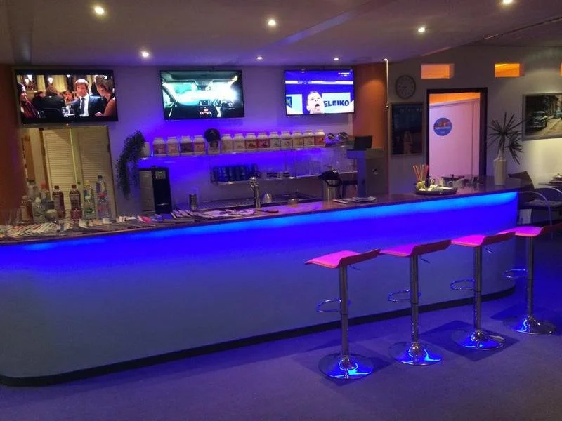 Ein Barbereich mit blauer LED-Beleuchtung, vier pinkfarbene Barhocker, mehreren Fernsehern an der Wand, Getränken auf der Bar, und einer modernen, stimmungsvollen Atmosphäre.