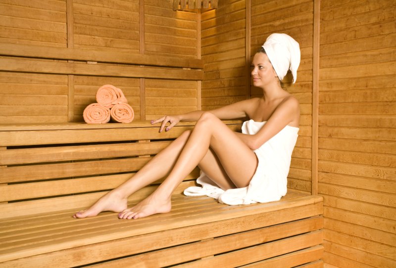 Eine junge Frau sitzt in einer Sauna, eingewickelt in ein Handtuch, mit einem weiteren Handtuch um den Kopf gewickelt. Es liegen drei weitere Handtücher auf einer Holzbank hinter ihr.