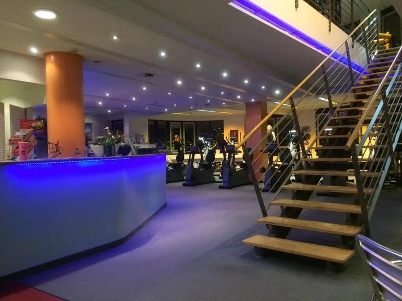 Innenansicht eines modernen Fitnessstudios mit Trainingsgeräten, einer Treppe und blauem Licht.
