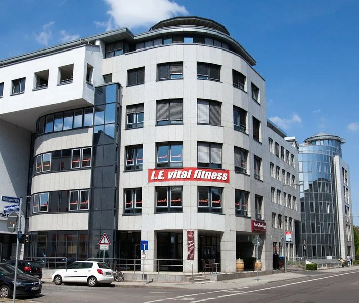 Modernes graues Bürogebäude mit Fenstern und einem Fitnessstudio-Schild in der Mitte, umgeben von Straßen und Autos.