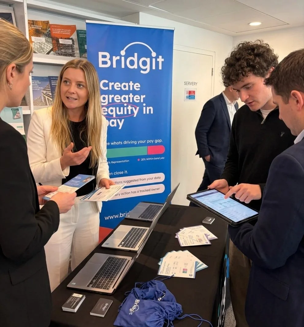 People gathered at a Bridgit booth discussing pay equity, with laptops, tablets, brochures, and branded promotional materials.