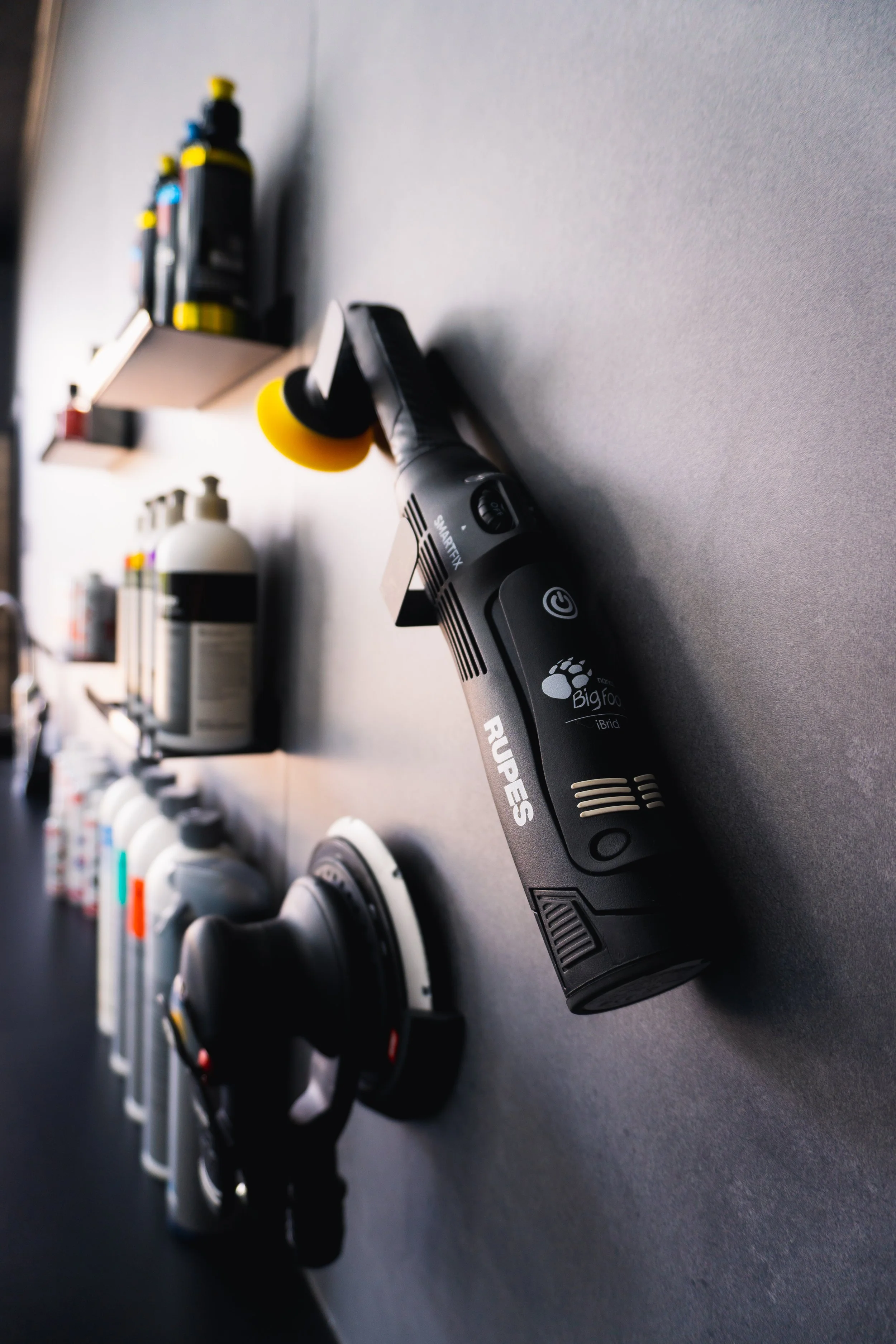 A black handheld device labeled 'RUPES' with buttons, resting on a gray surface. In the background, various bottles and containers are on shelves, including a white bottle with a pump and smaller bottles with colorful caps.