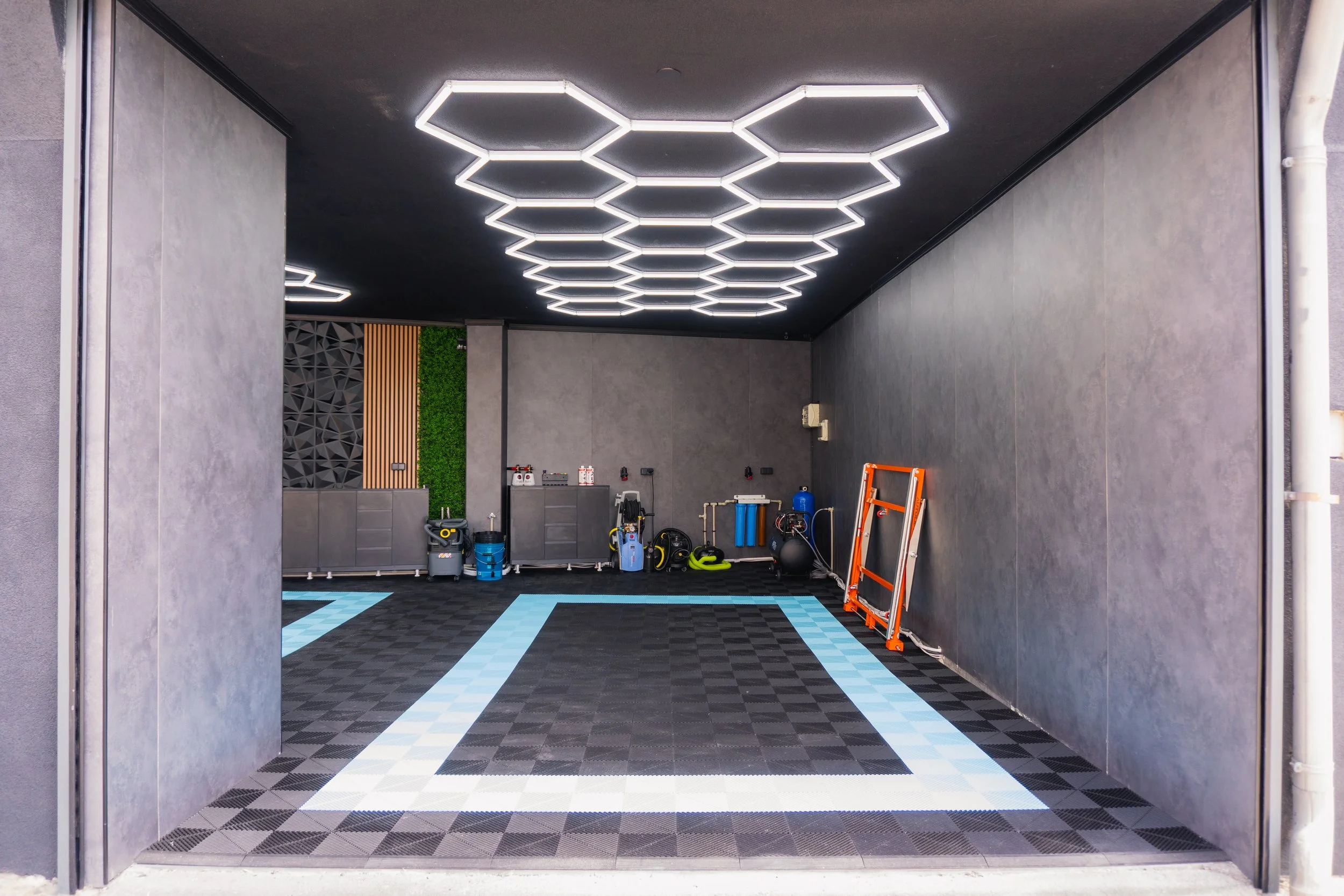 View of a modern gym or fitness room with hexagonal LED ceiling lights, black safety mats, exercise equipment, and a wooden and green wall accent.