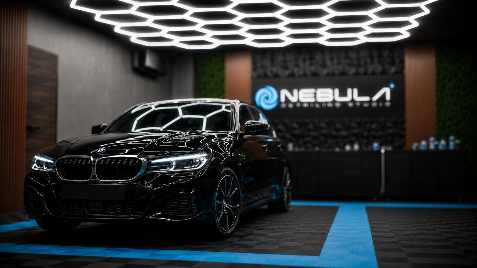 A black BMW parked inside a modern, illuminated garage with a glowing ceiling design. The garage features a sign that says 'Nebula' with a swirling logo in the background.