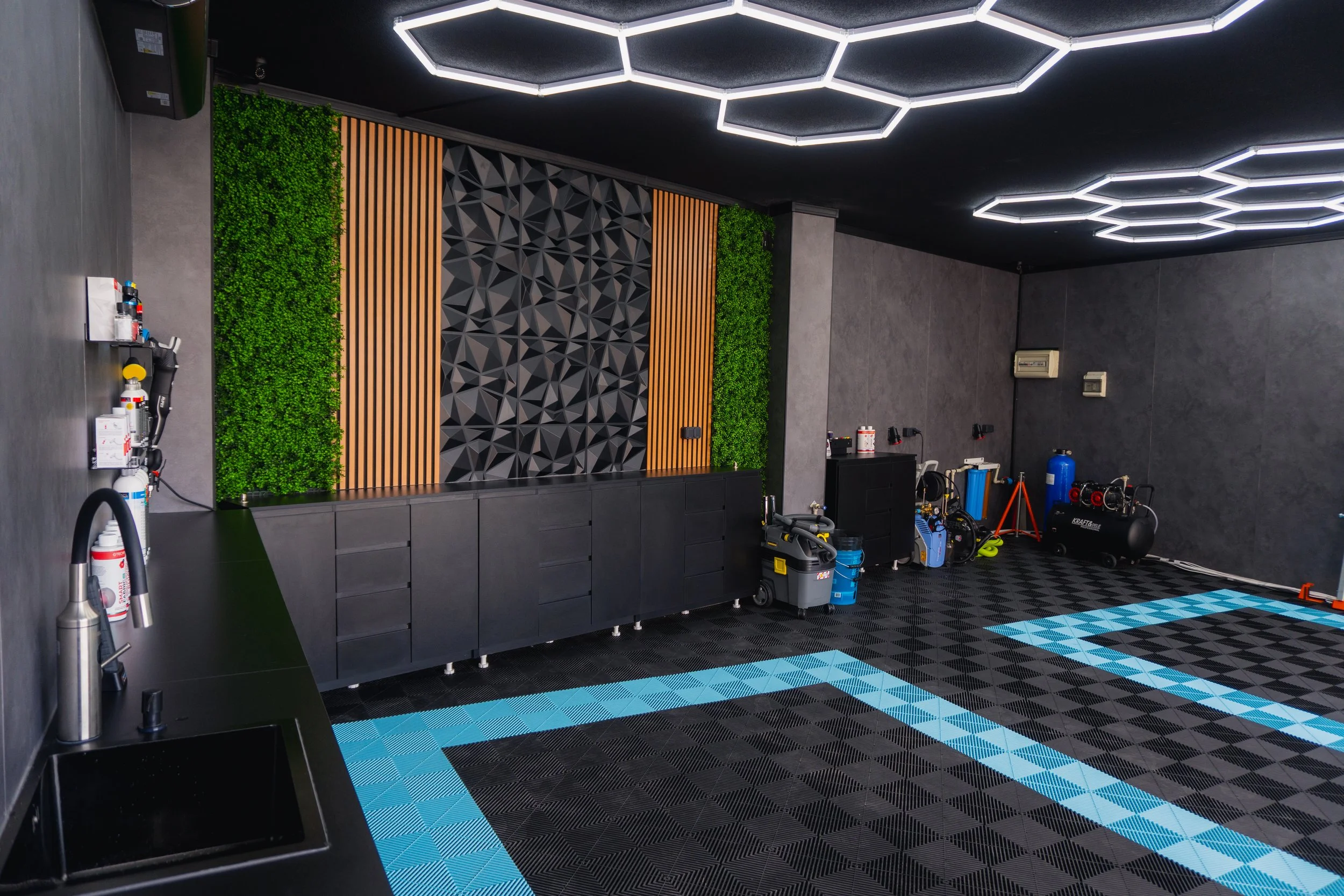 A modern interior gym space with black and gray walls, a black and blue checkered rubber flooring, and wall-mounted greenery and wood panels. Equipment such as fire extinguishers, a black cabinet, and various gym tools and machines are visible, along with ceiling hexagonal LED lights.