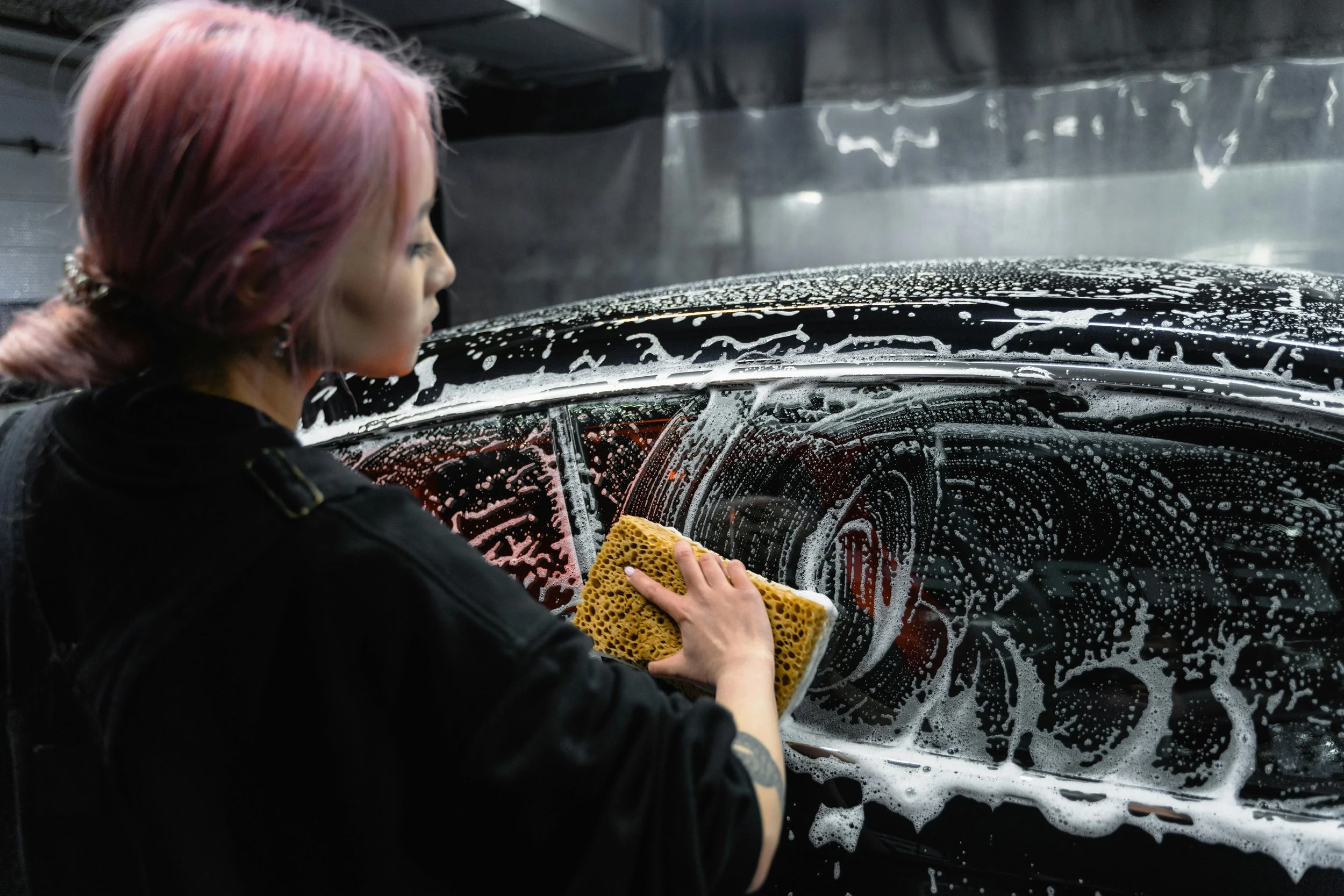 A woman with pink hair washing a black car with soap and a yellow sponge.