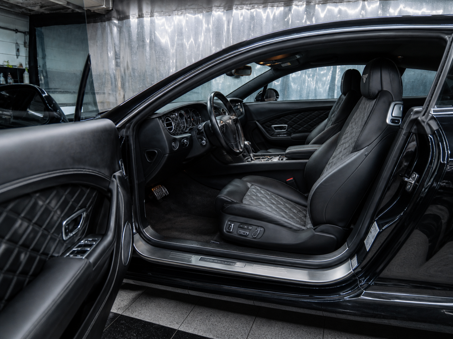 Interior of a black luxury sports car, showing the driver's seat, steering wheel, dashboard, and part of the passenger seat, with quilted leather upholstery and various controls.