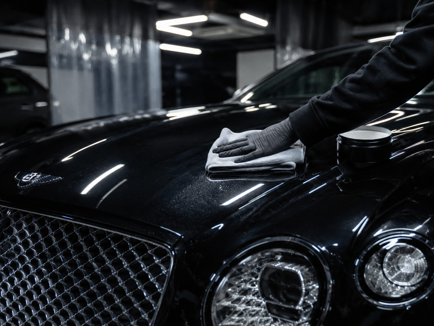 Person in black clothing and gloves cleaning a black luxury car with a cloth in a garage.