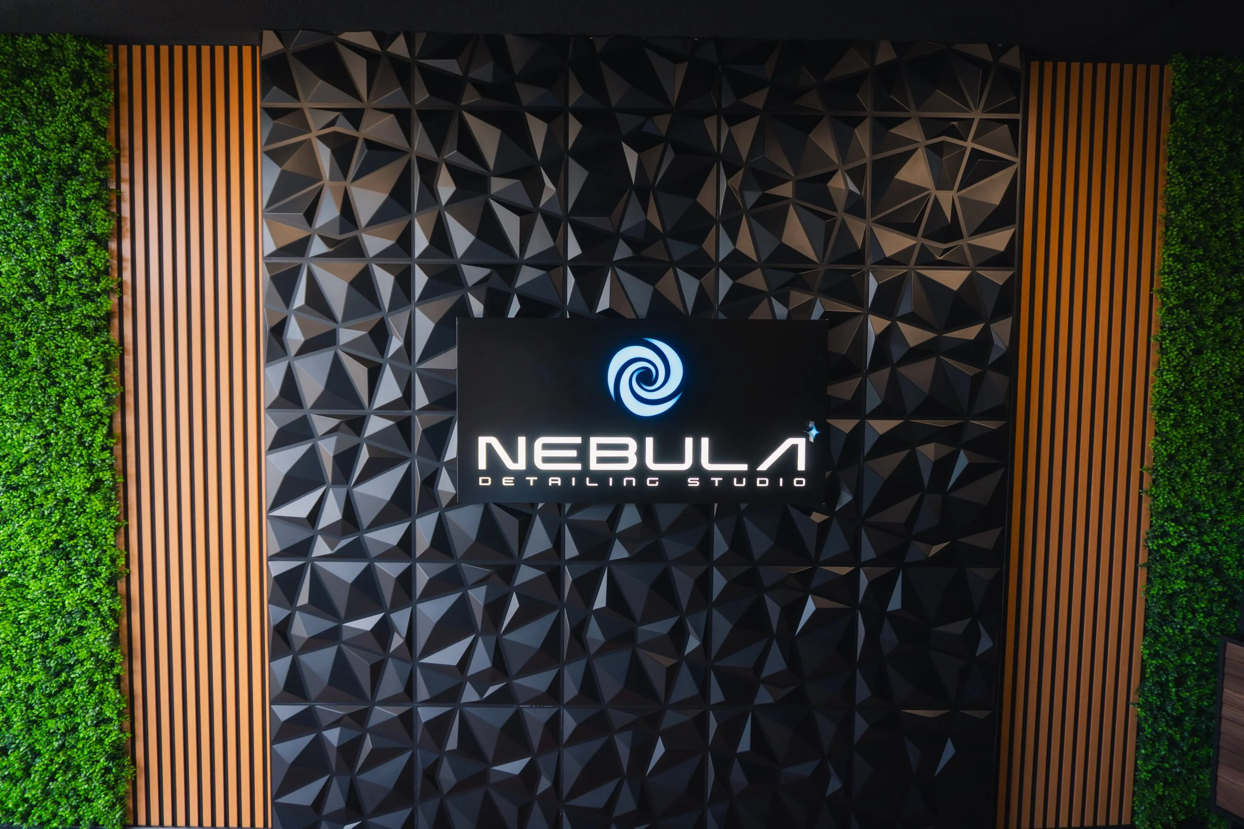Interior view of a modern design studio with a textured black wall, wooden slats on either side, green foliage on the edges, and a illuminated logo with the text 'Nebula Detailing Studio' in the center.