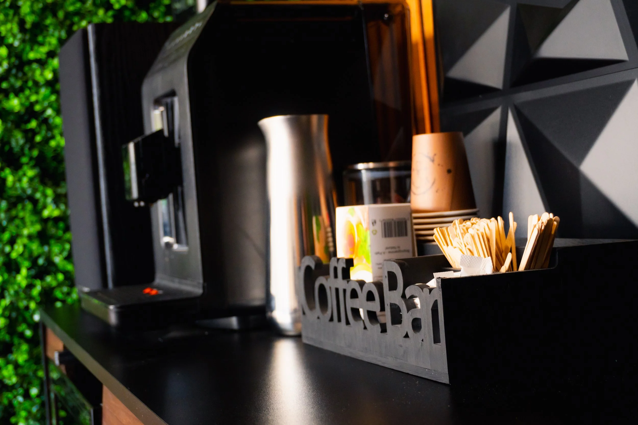 A coffee station with a black countertop, a silver thermos, a carton of juice, a stack of paper cups, and a container of wooden stir sticks, set against a geometric-patterned wall and green foliage background.