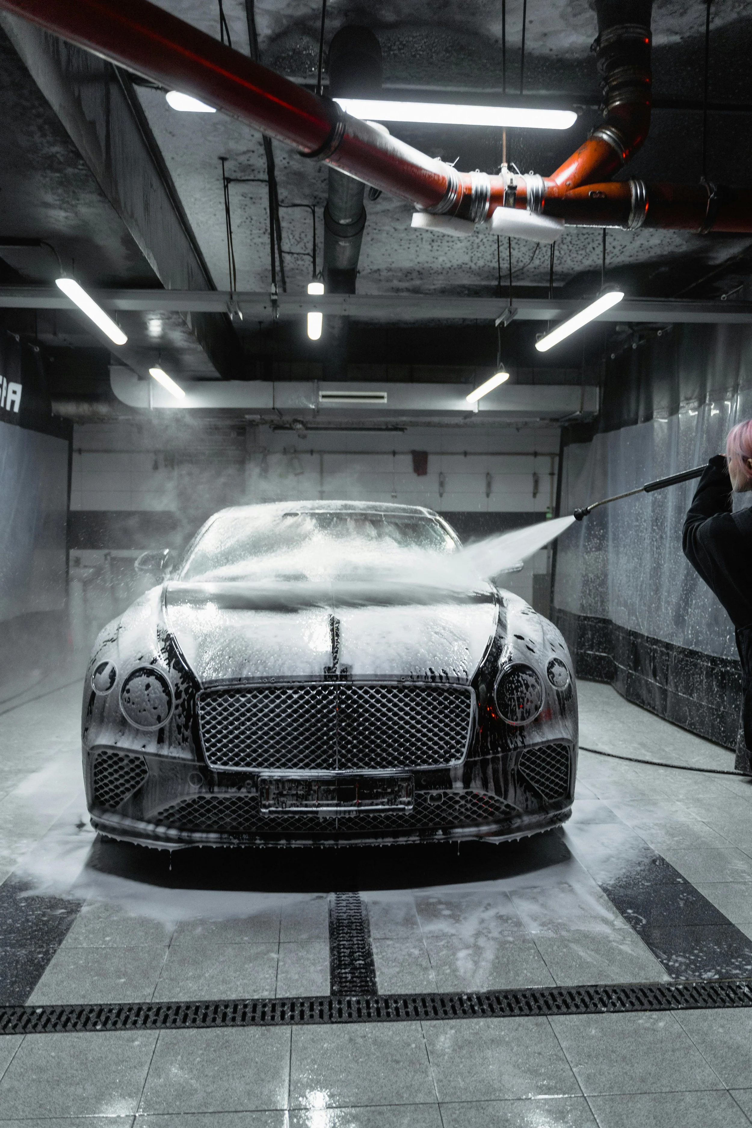 A black luxury car being washed in an industrial-style car wash with overhead fluorescent lights and red and black pipes on the ceiling. A person with pink hair operates a high-pressure water hose on the car.