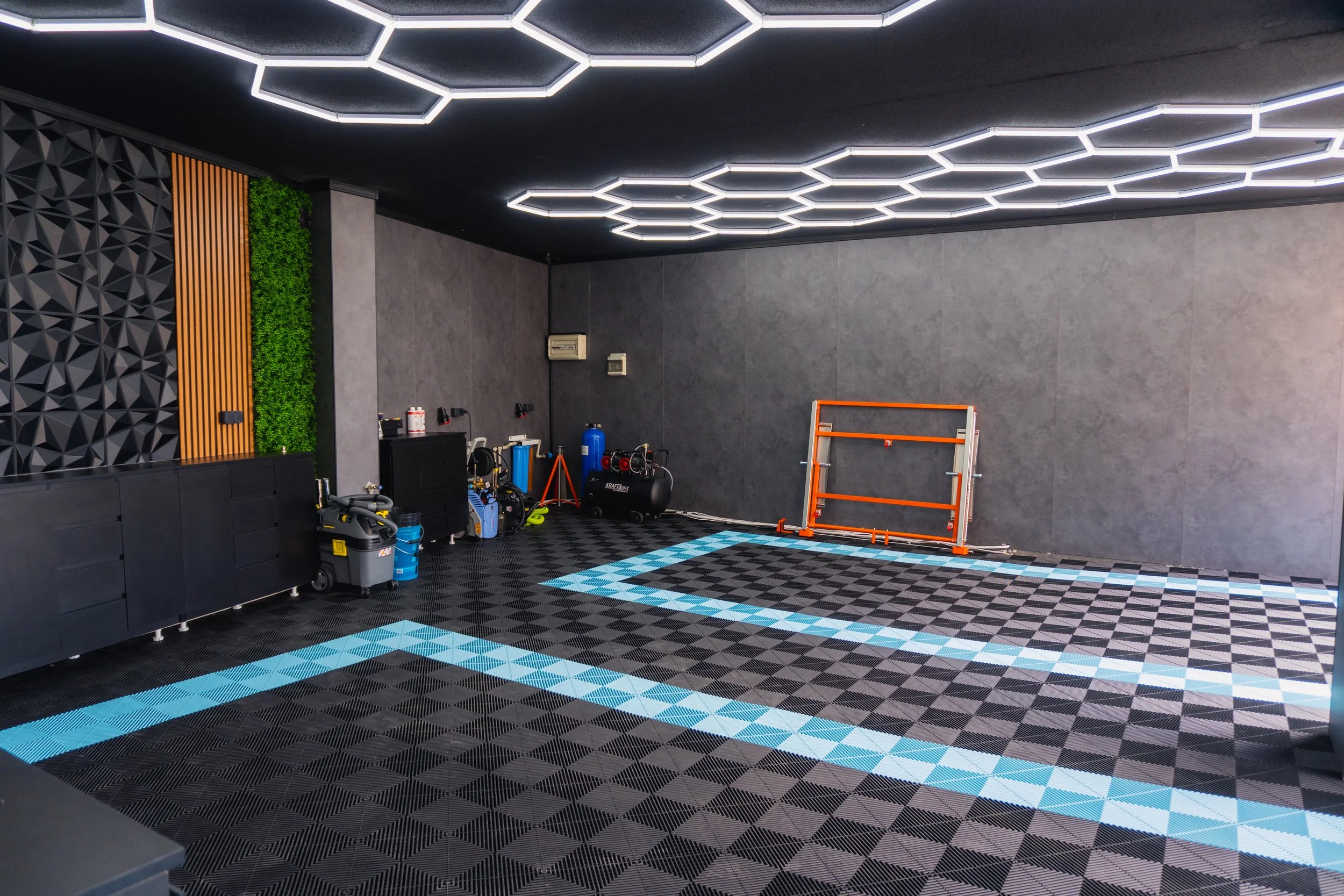 A modern interior garage or workshop with hexagon-shaped LED light fixtures on a black ceiling, black and gray walls, and a multi-colored patterned rubber floor. Various tools, equipment, and a safety frame are arranged along the walls.