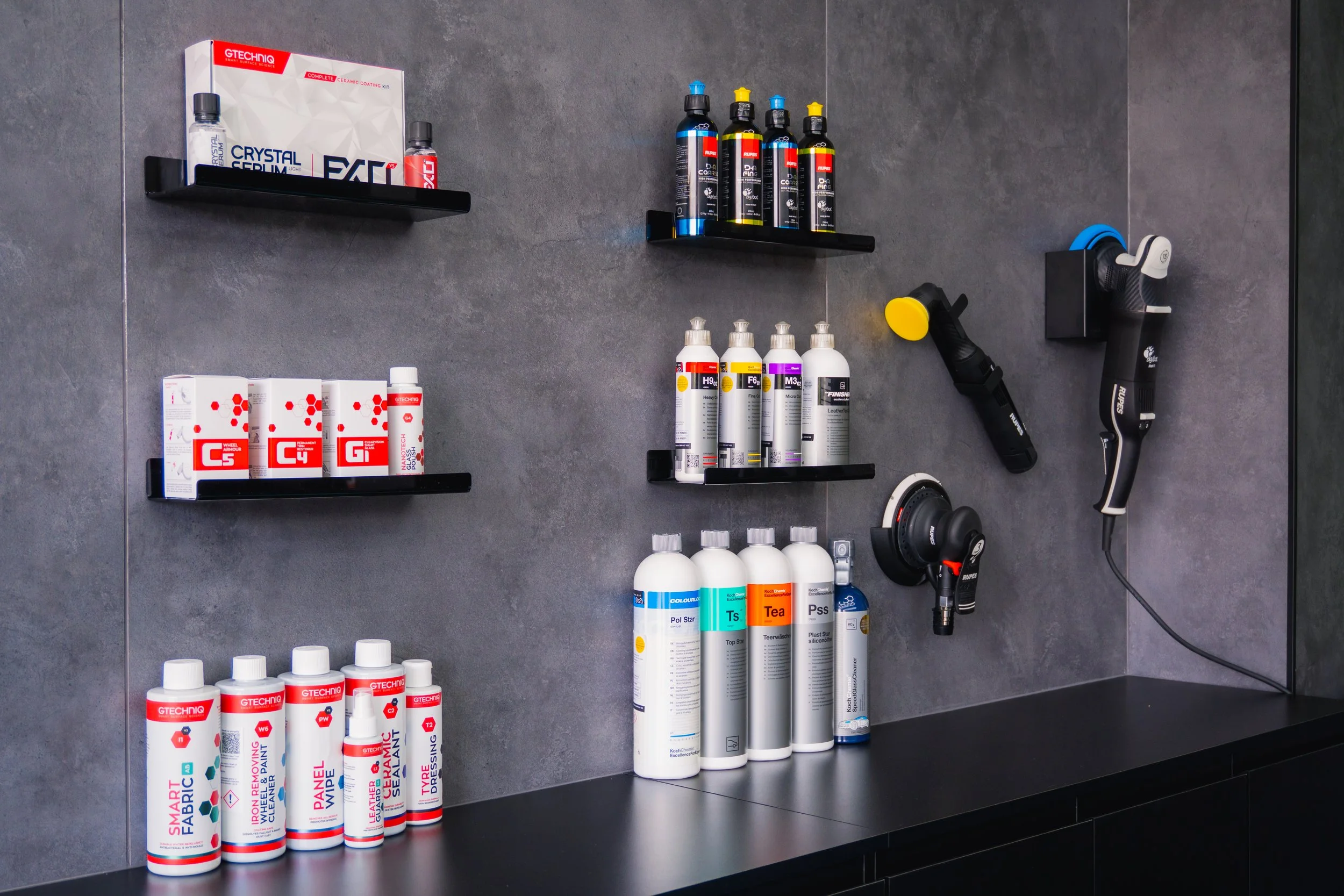 A wall-mounted display shelf with various automotive and repair products including sprays, cans, and cleaning solutions. Items are arranged on black shelves against a gray tiled wall, with tools and equipment hanging nearby.
