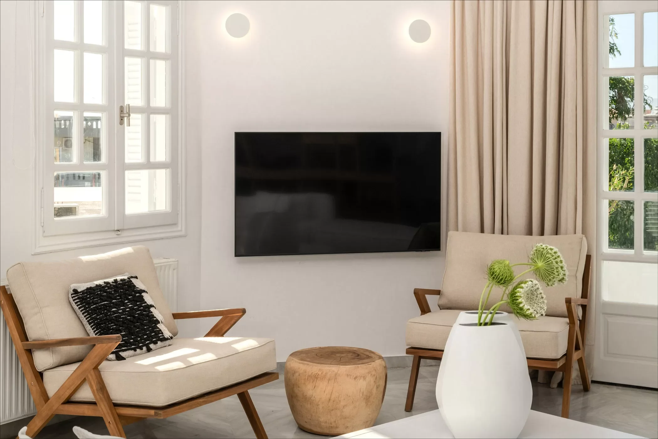 Living room with beige sofas, a wooden stool, a large white vase with green flowers, a mounted TV, and windows with beige curtains.