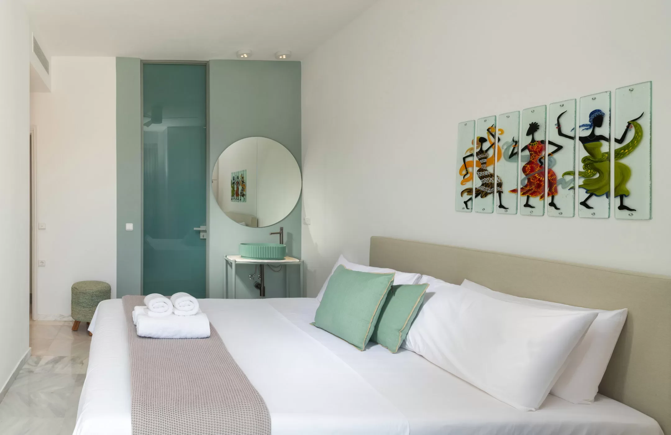 A bedroom with a king-sized bed decorated with white sheets, two green pillows, and a beige bed runner. A set of white towels is placed at the foot of the bed. On the wall, above the bed, is a colorful African-style art piece depicting dancing figure