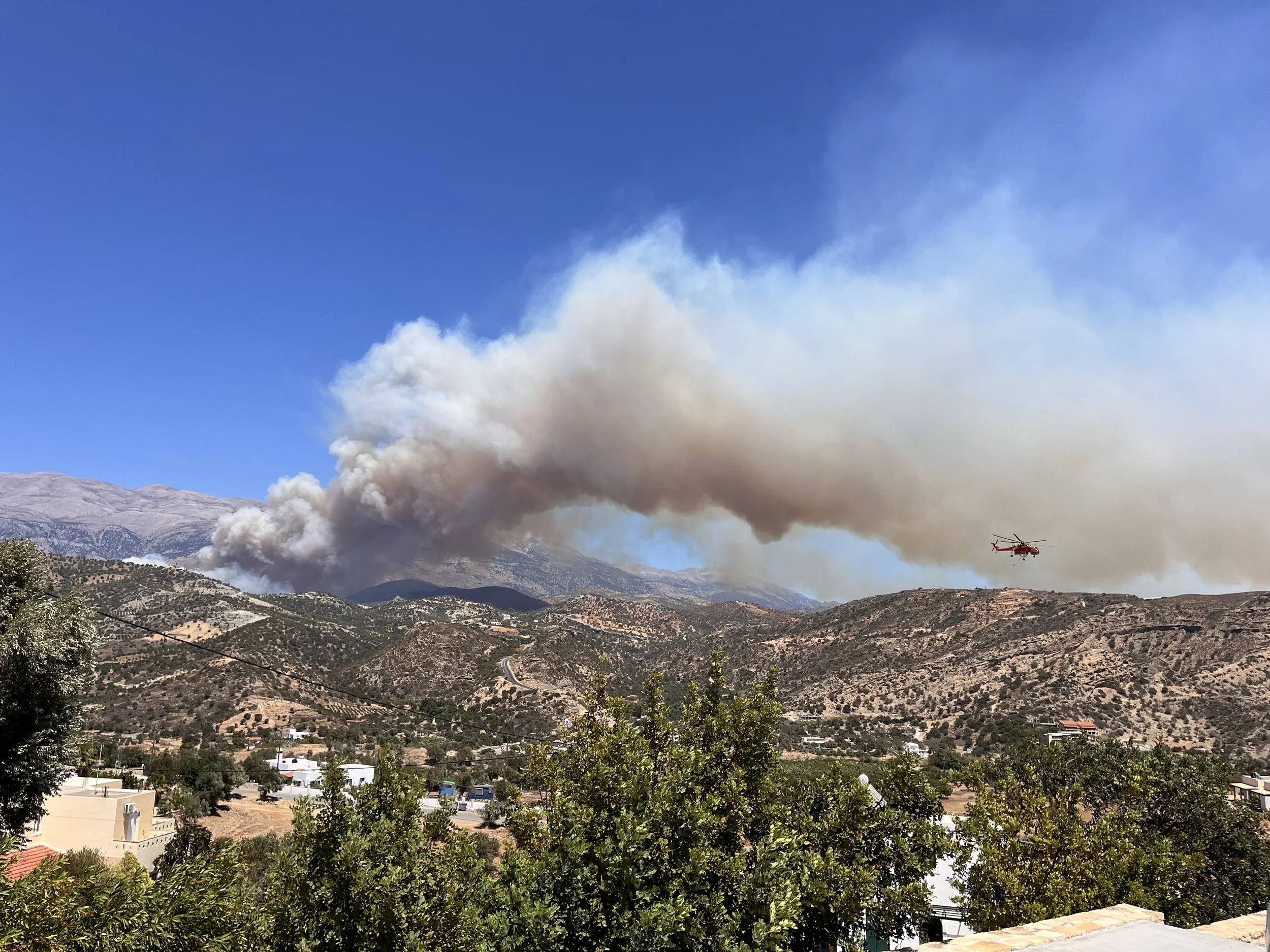 Smoke plume from the Amari Valley wildfire rising above the hills of South Crete as a firefighting helicopter approaches the fire zone.