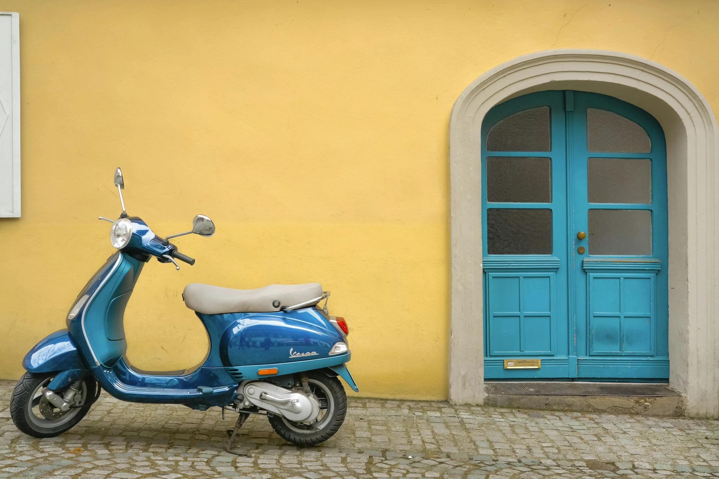 Blue scooter parked on cobblestone street in front of a yellow wall with a blue arched door. Scooter Rental Crete Greece near Matala and Triopetra