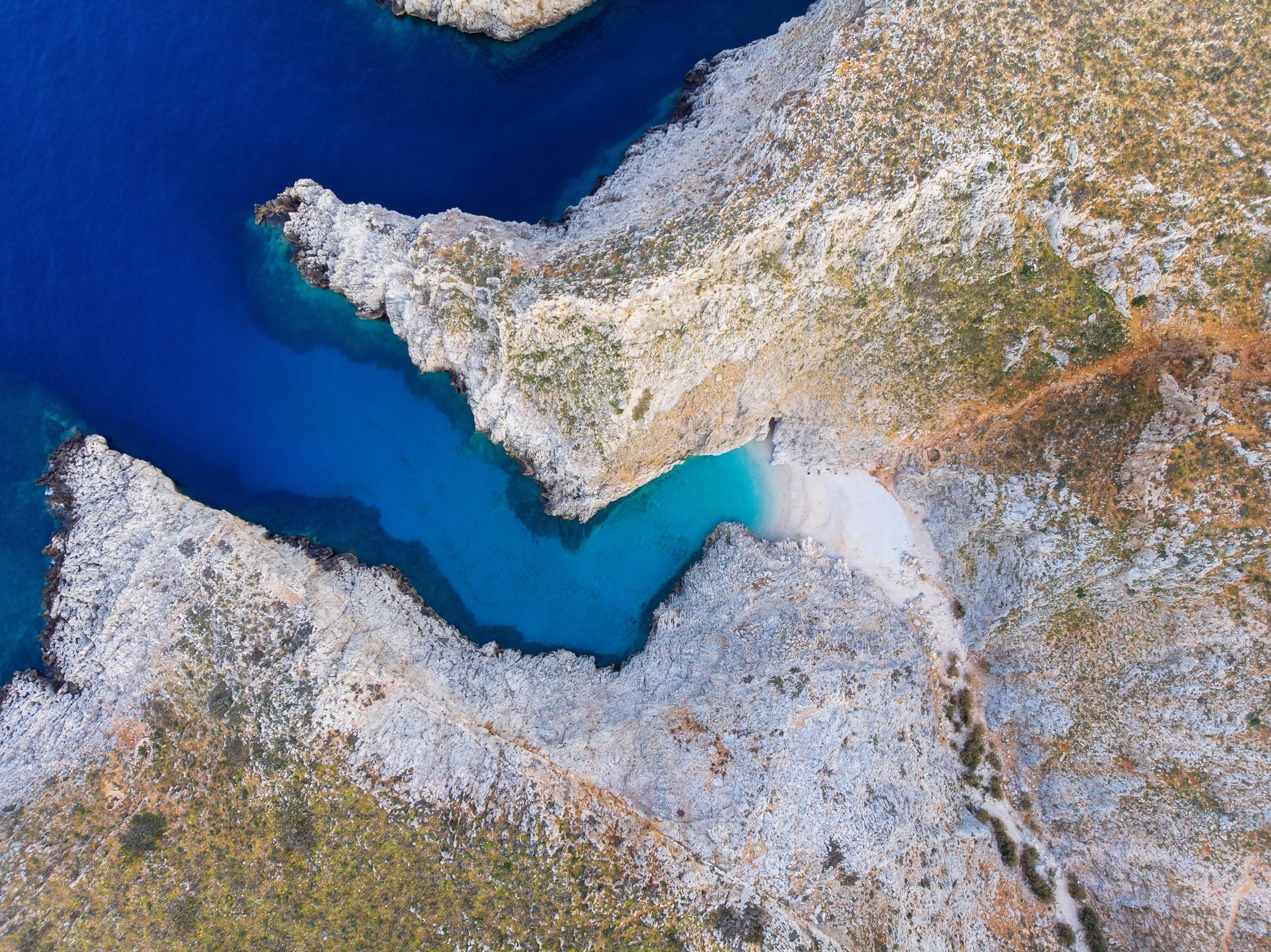 Aerial image of a rugged coast in Crete, with blue and turquoise water and a sandy cove.