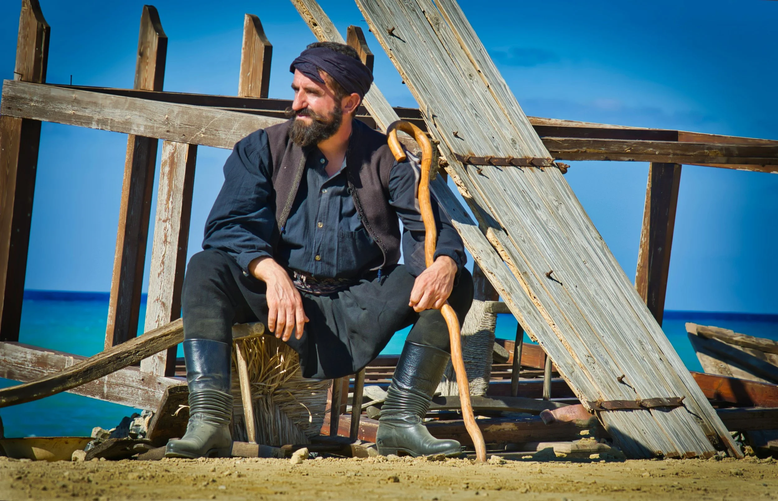 A man with a beard and headscarf sitting outdoors on a sandy surface near a partially collapsed wooden structure, holding a walking stick, with an ocean and blue sky in the background.