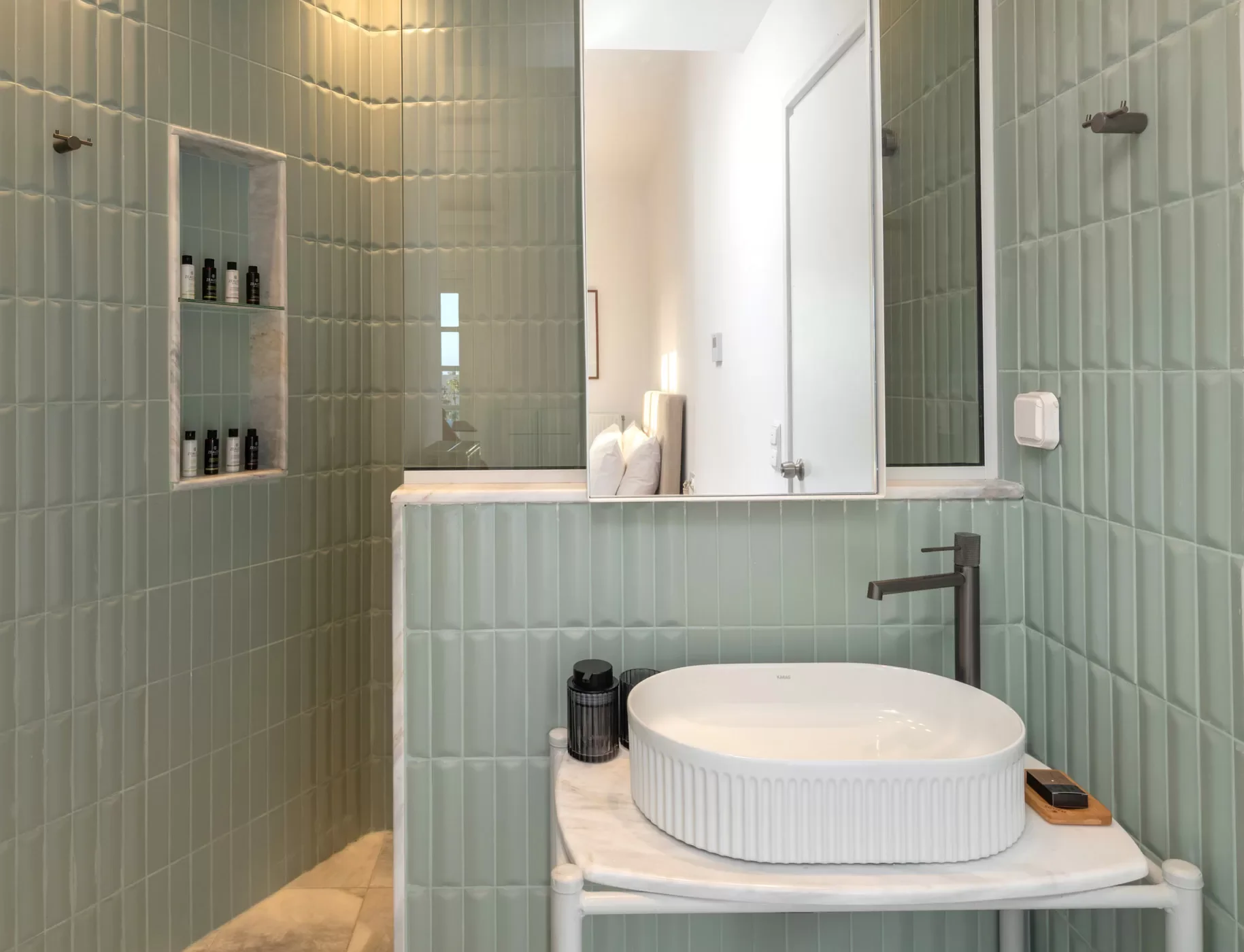 Modern bathroom featuring green tiled walls, a round vessel sink on a white marble countertop, with a black faucet, a mirror, and built-in shelves with toiletries