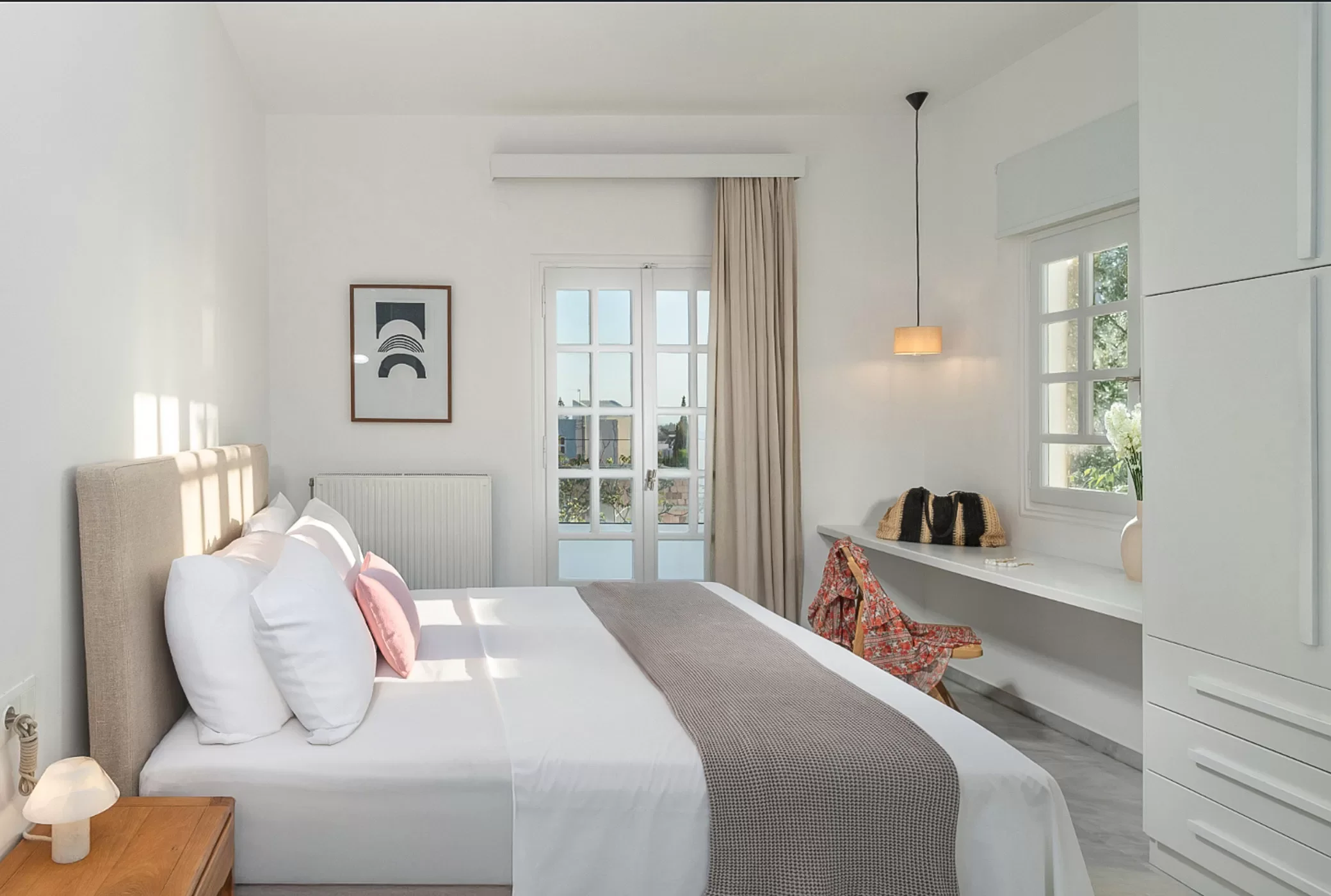 A bright, minimalist bedroom with a bed, bedside table, and a window showing trees outside.