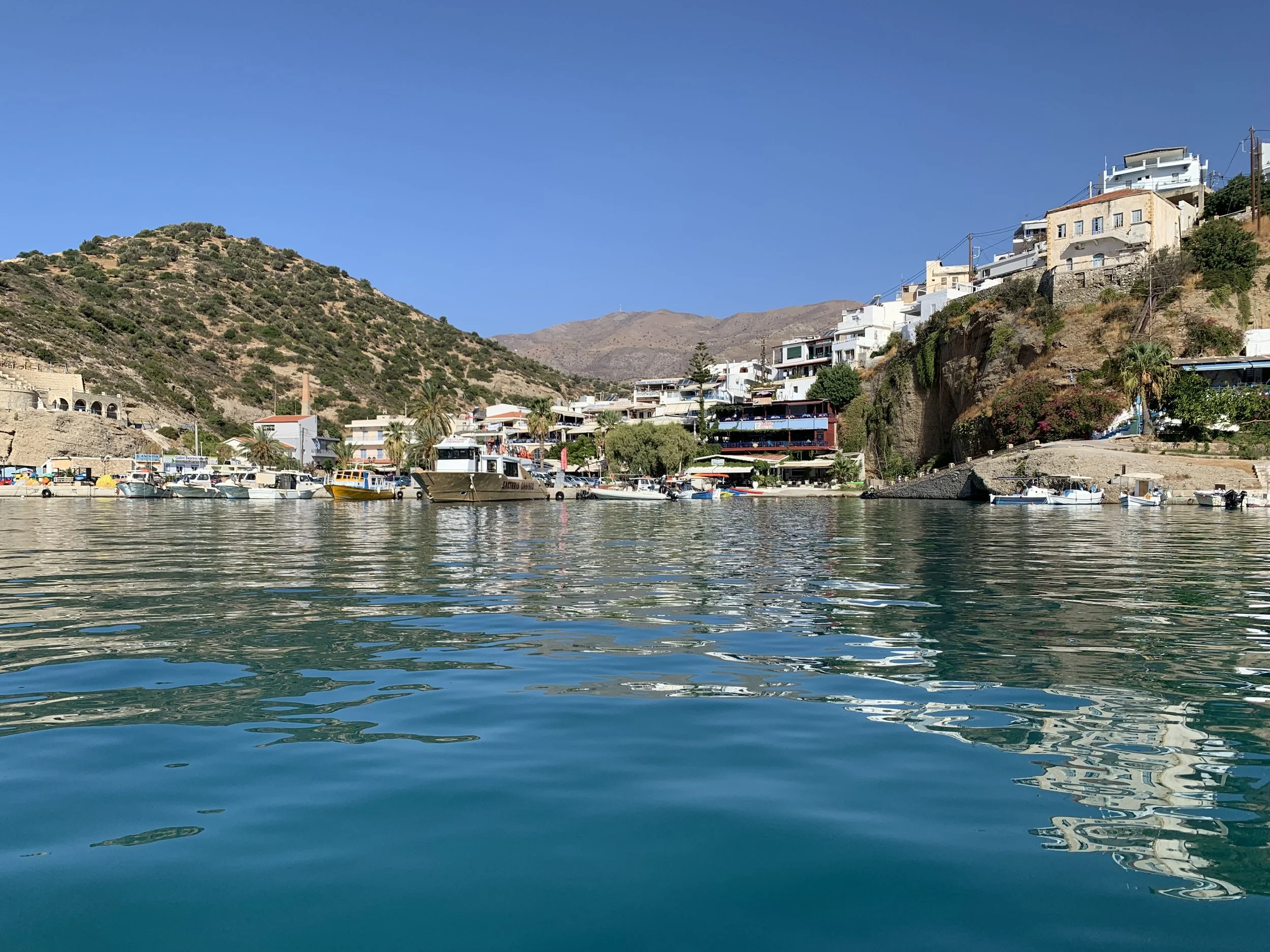 View of Agia Galini harbour from the sea, with fishing boats, waterfront tavernas and whitewashed houses climbing the hillside above the bay.
