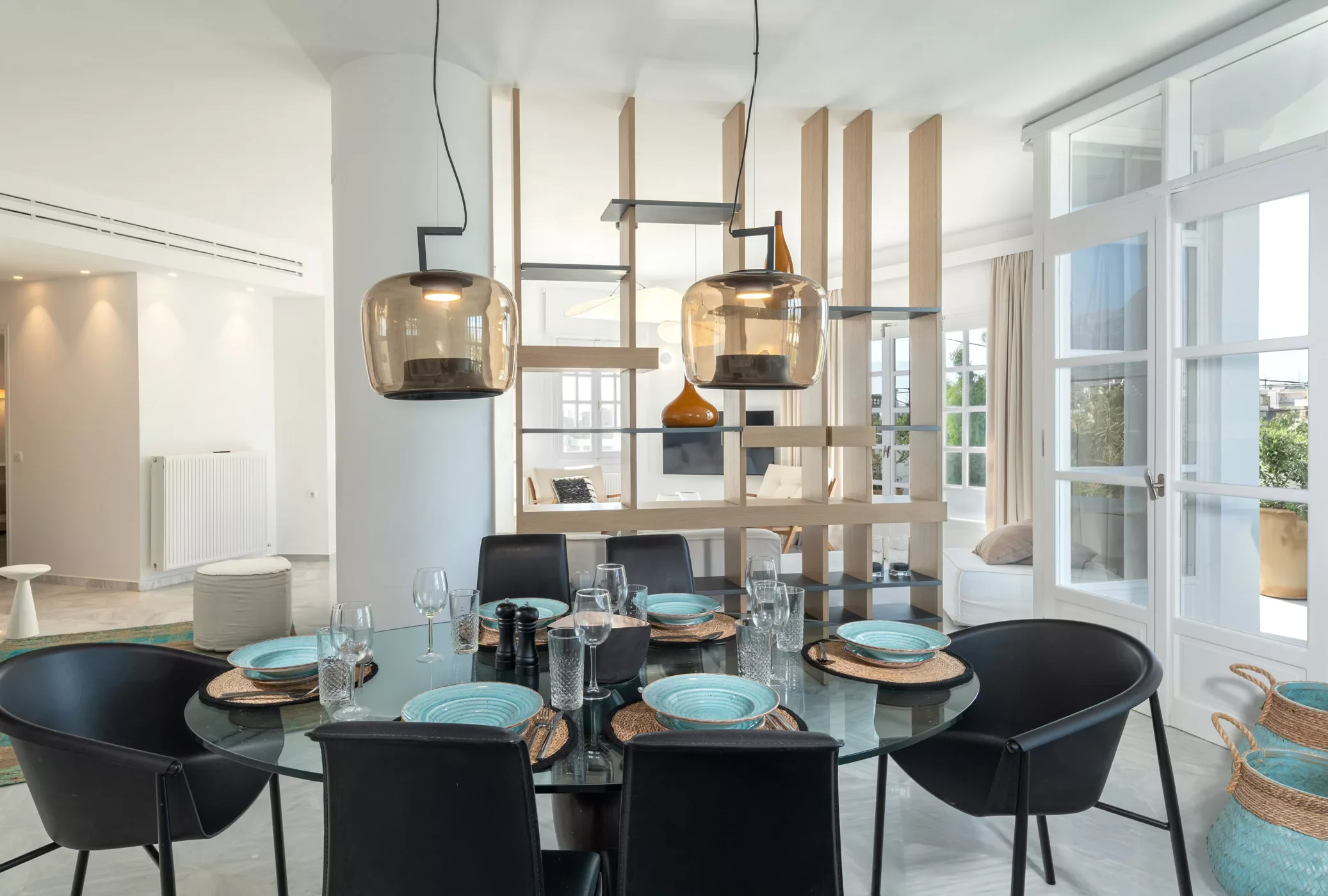 Modern dining room with a round glass table set with turquoise plates, black bowls, and clear glasses, surrounded by black chairs. Two pendant lights hang above the table. Behind, a decorative wooden partition separates the dining area from a living 