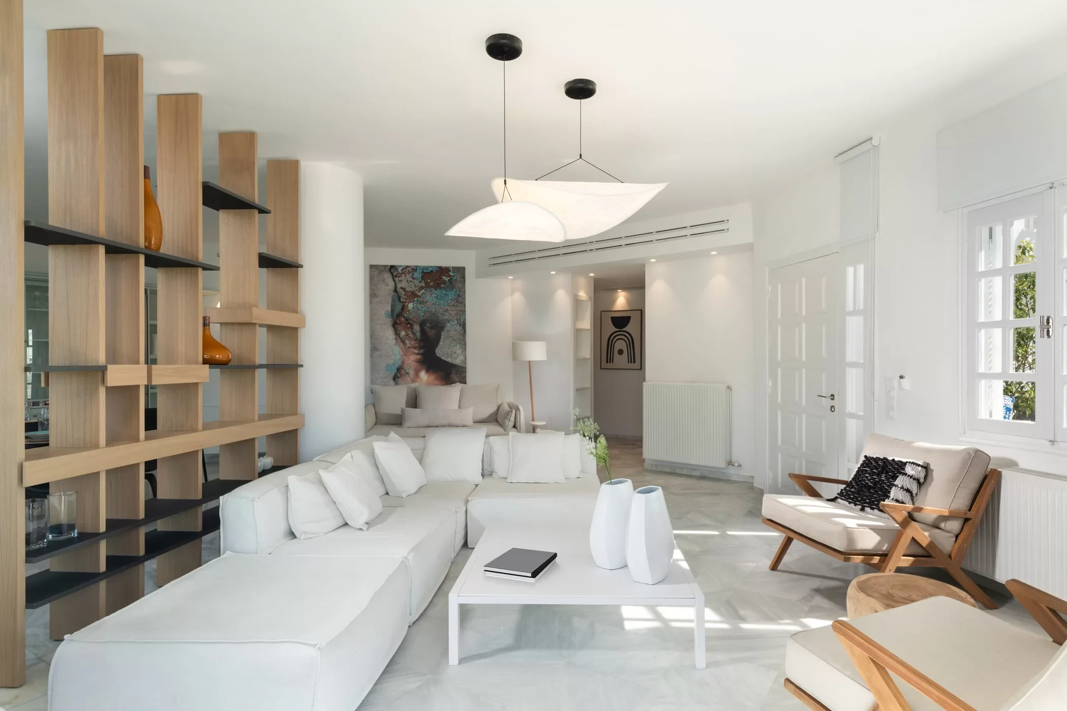 Bright living room with white sofa, beige armchair, wooden and black shelving, white coffee table, modern art on white walls, and large windows letting in natural light.