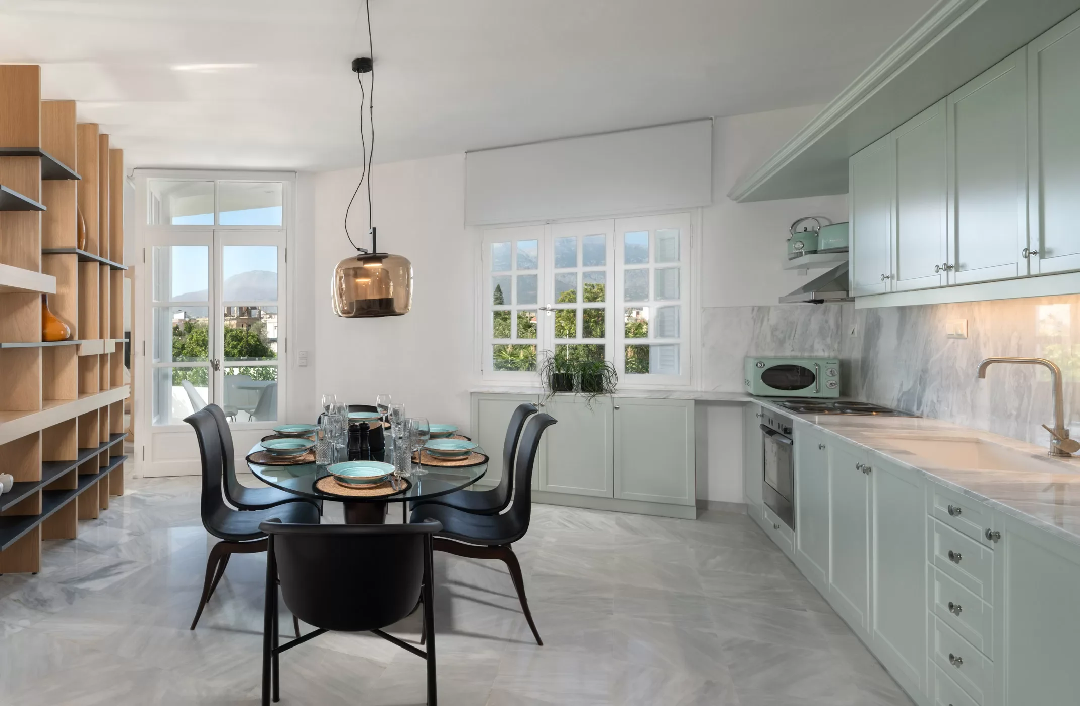 Modern kitchen and dining area with a round black table set with plates, glasses, and utensils, surrounded by black chairs, with large windows showing a view of trees and mountains, white cabinets, marble countertops, and a wooden shelf on the left.