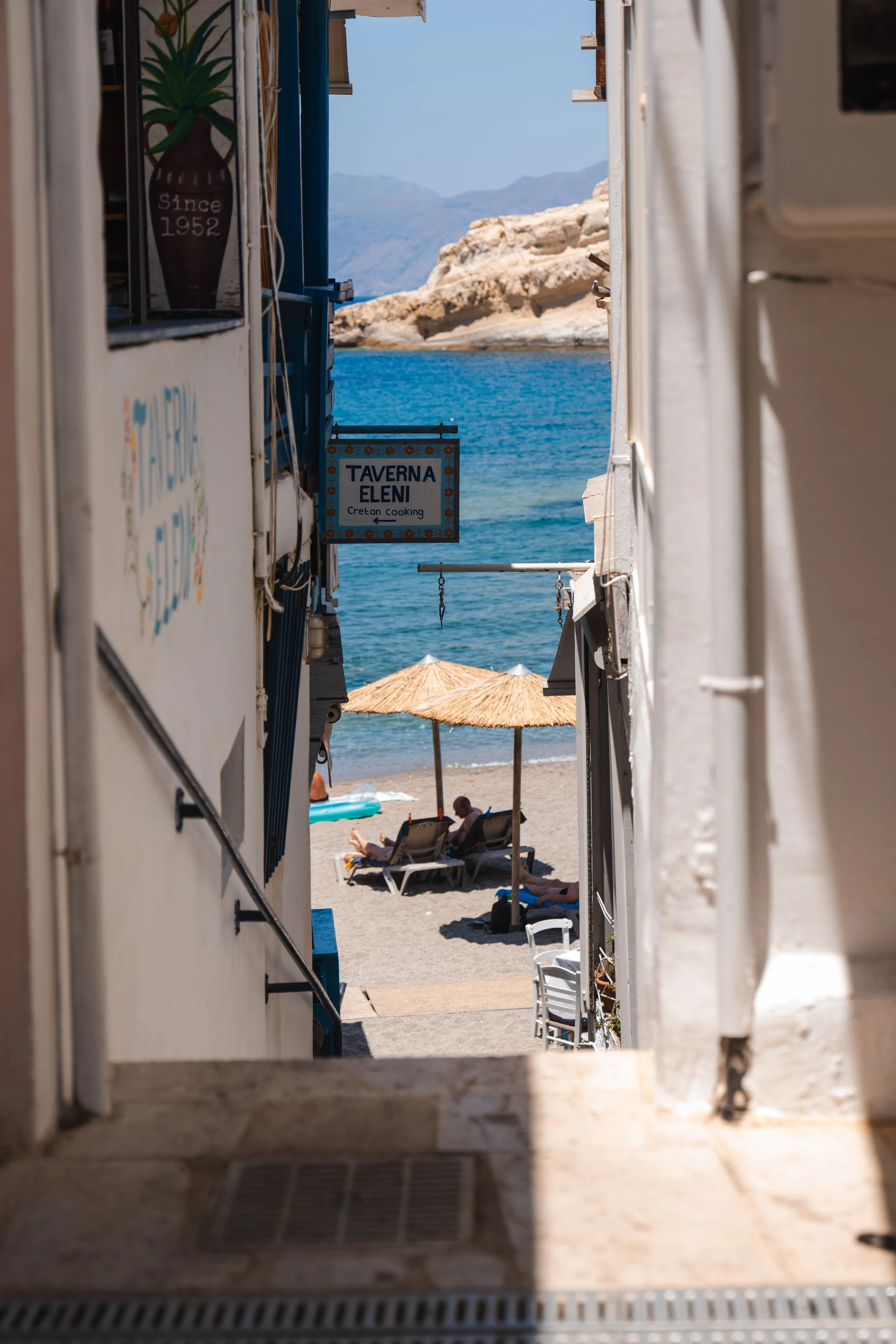 Facades of houses framing a view of umbrellas and the sea at Matala Beach, Crete.