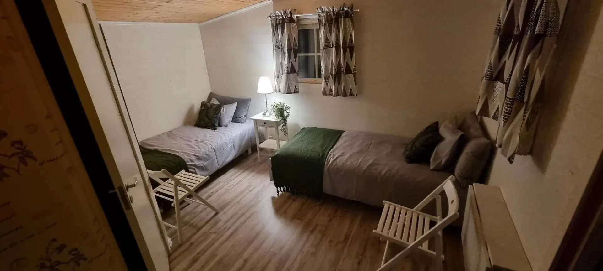 Ein Schlafzimmer mit zwei Einzelbetten, einem Nachttisch mit Lampe und einer Pflanze, einer Fenster mit patterned Vorhängen, einem Holzfußboden und minimaler Einrichtung.