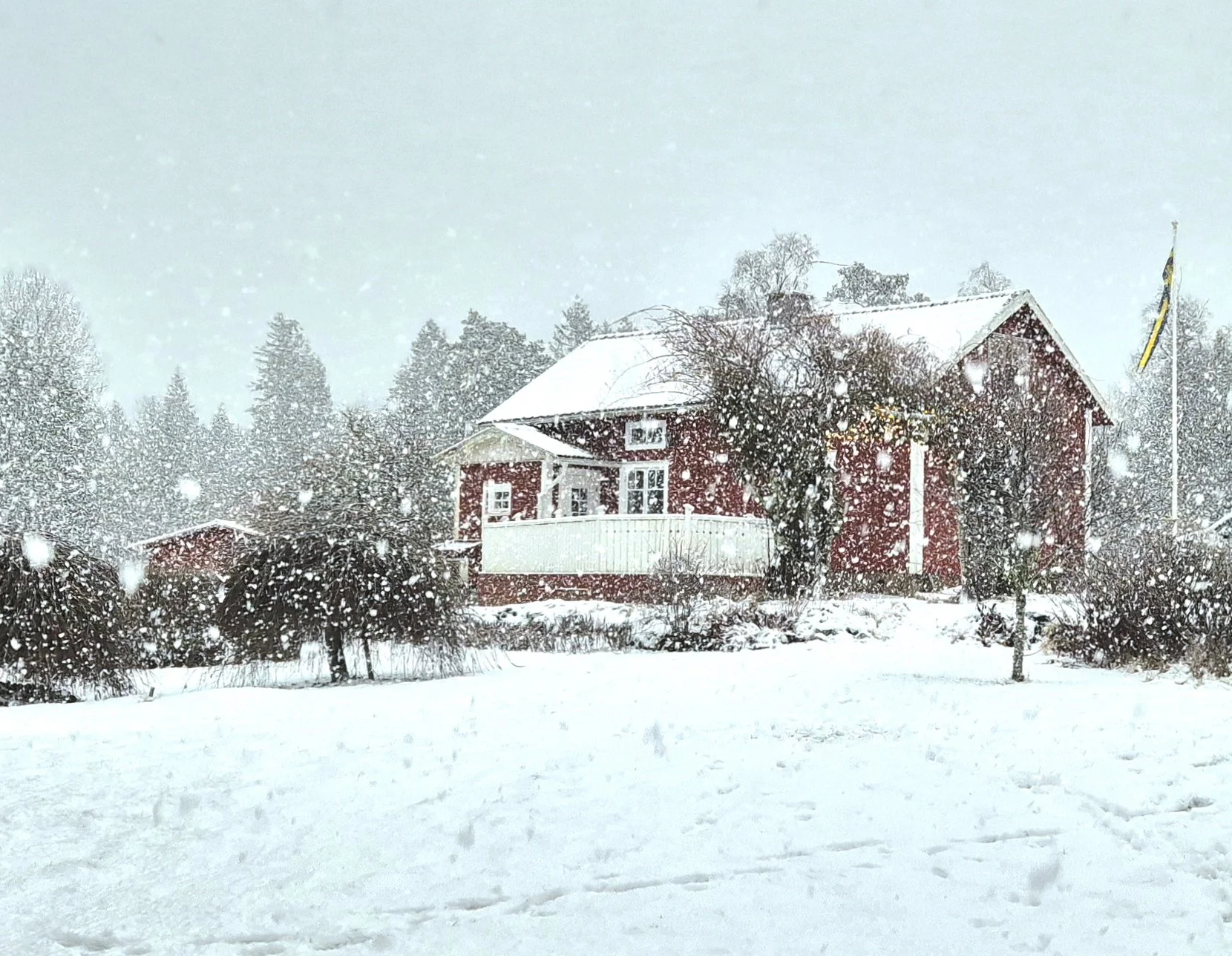 Ein verschneites Haus in einer Winterlandschaft mit Schnee bedecktem Boden, Bäumen und einem schneebedeckten Dach, während Schneeflocken fallen.