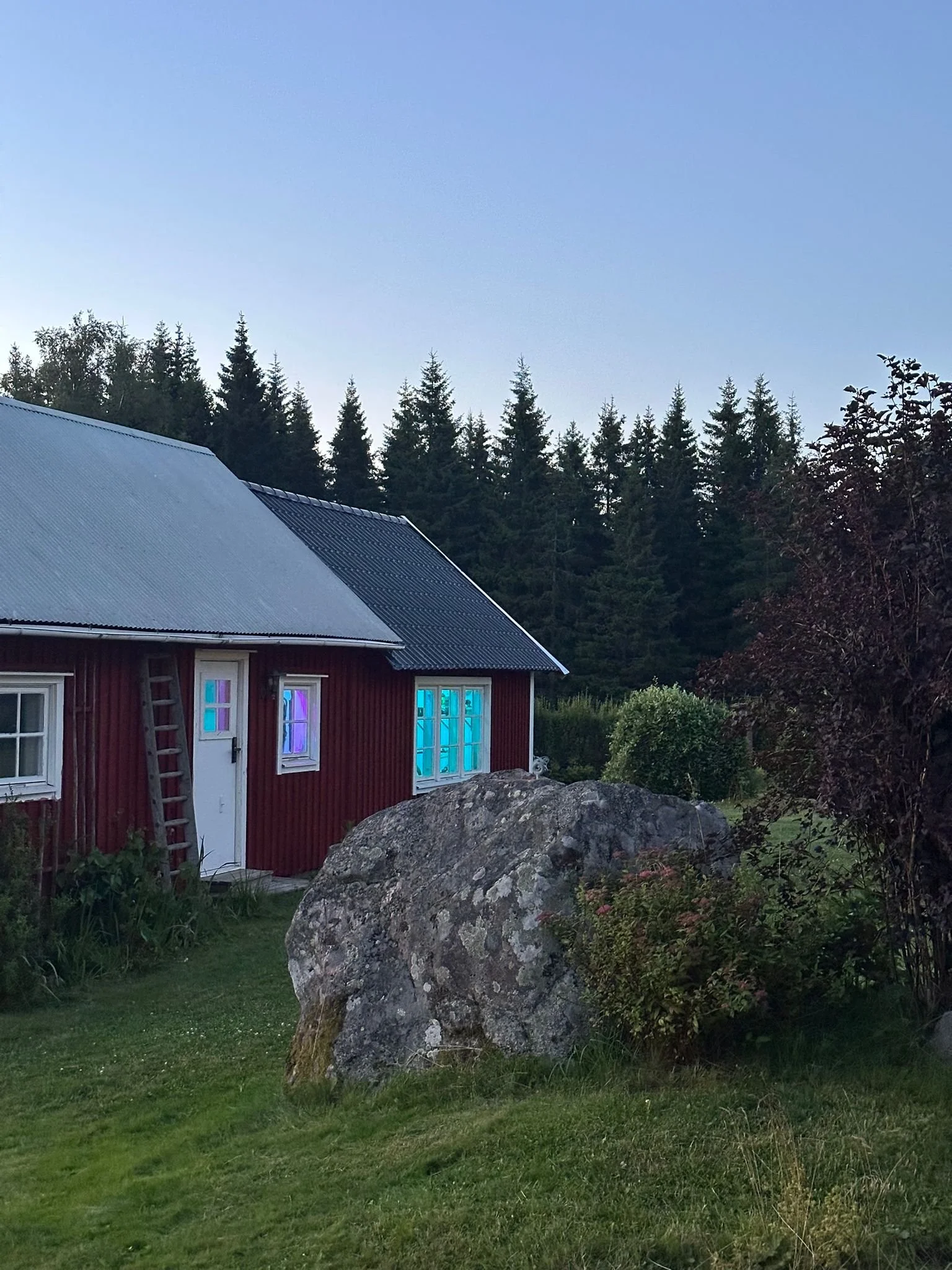 Ein rotes Holzhaus mit blauen, bunten Fenstern vor einem grünen Garten mit großen Steinen und einem Nadelwald im Hintergrund bei Dämmerung.