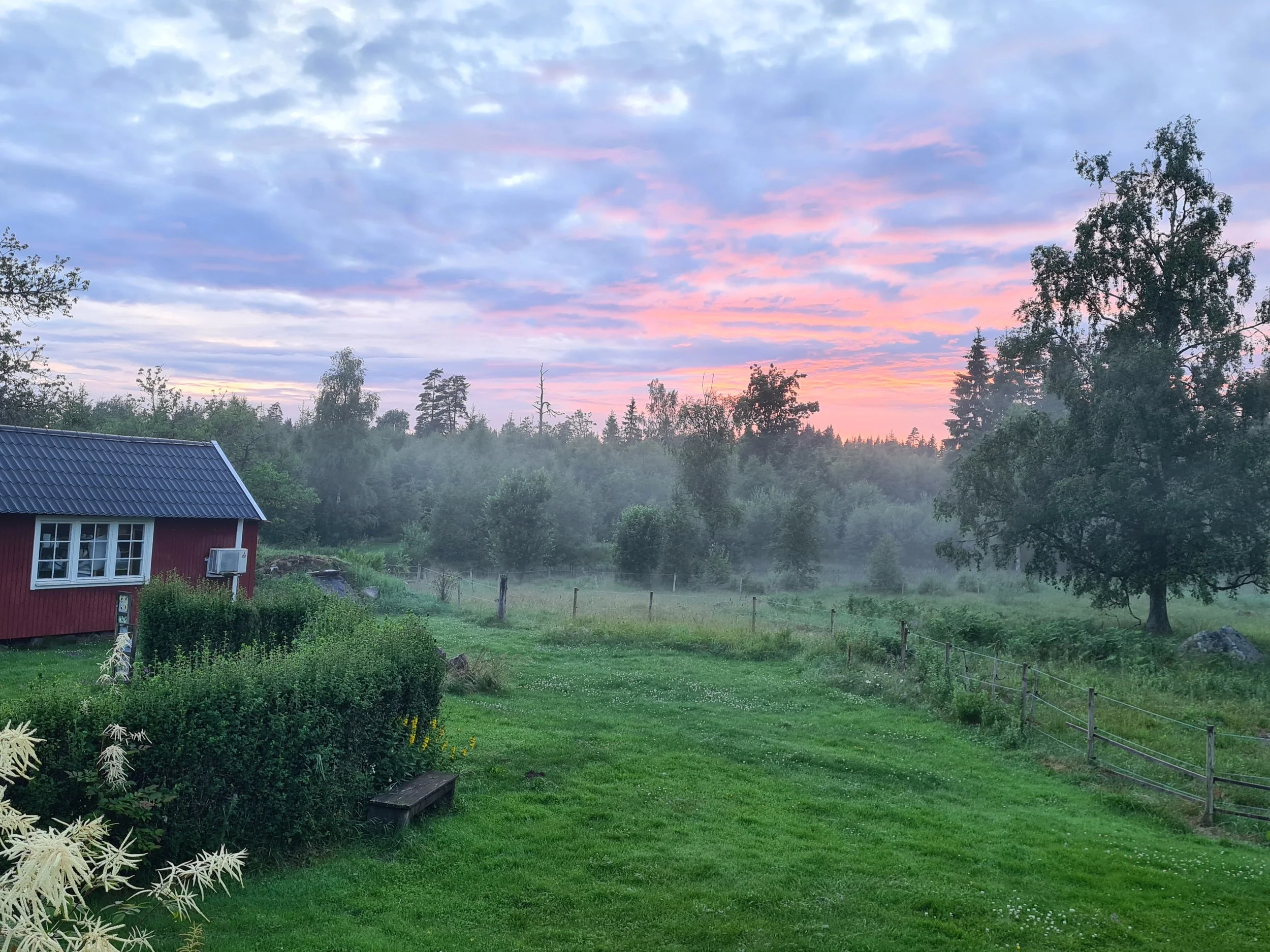 Ein ländliches Landschaftsbild bei Sonnenaufgang mit grünem Rasen, Bäumen, einer roten Holzhütte mit blauer Dachziegel, und einem Zaun, im Hintergrund ein bewölkter Himmel mit pastellfarbenem Sonnenaufgang.