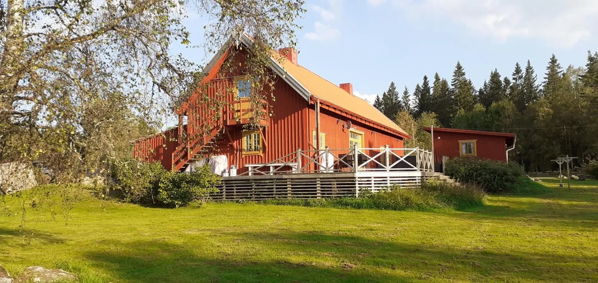 Ein rotes Holzhaus auf einer grünen Wiese mit Bäumen im Hintergrund, Sonnenschein und blauem Himmel.