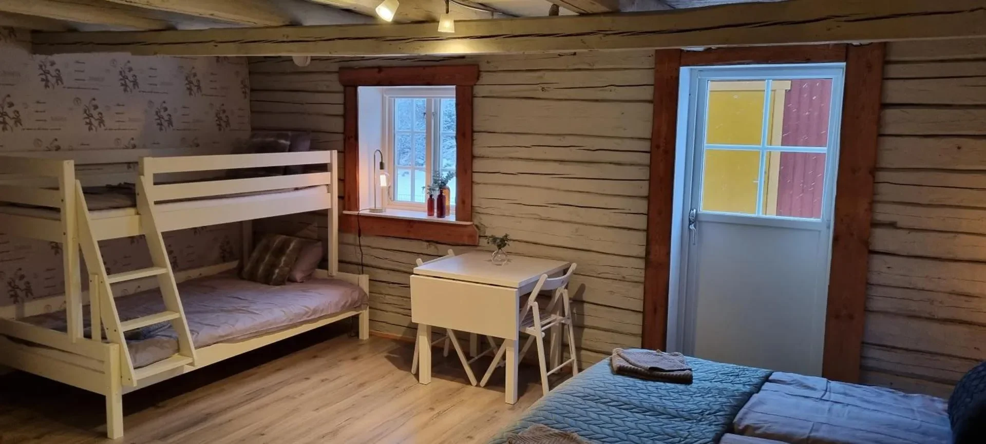 Ein Schlafzimmer mit Holzwänden, einem Doppelbett, einem Hochbett, einem kleinen Tisch mit Stühlen, einem Fenster und einer Tür. Die Einrichtung ist schlicht und gemütlich.