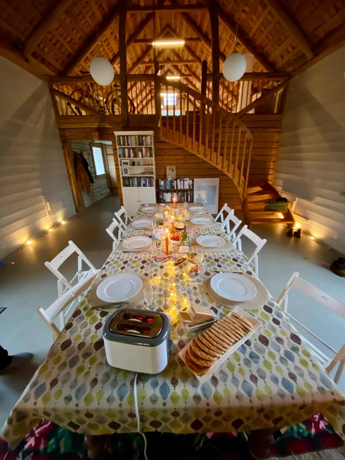 Ein festlich gedeckter Esstisch mit weißen Tellern, Besteck, Gläsern und Kerzen, im Hintergrund ein Holzregal mit Büchern, in einem Raum mit Holzdecke und Wänden, beleuchtet mit Deckenleuchten und Lichterkette auf dem Boden.