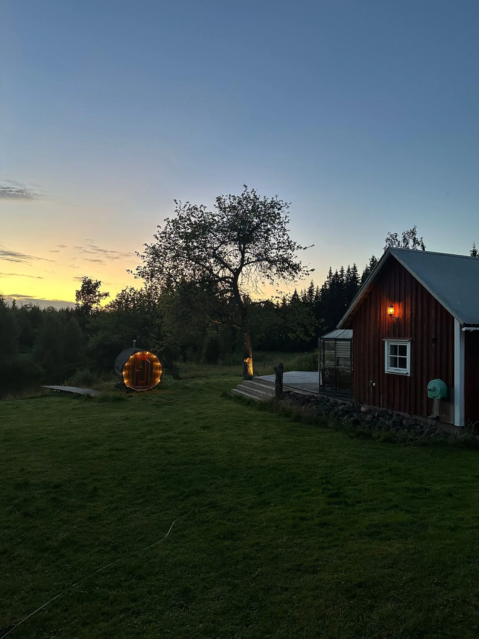 Sonnenuntergang über einer grünen Wiese mit einem Baum, einem roten Holzhaus mit beleuchtetem Außenlicht und einem runden, beleuchteten Zelt im Hintergrund.