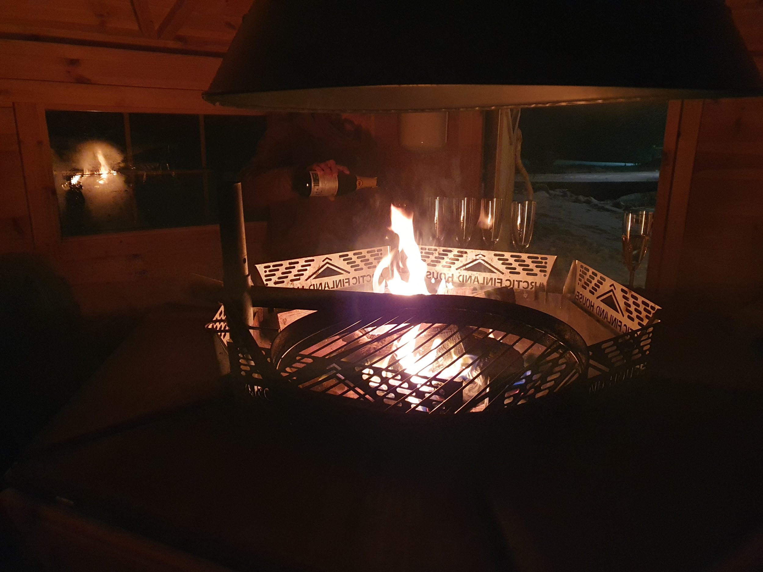 Ein Lagerfeuer in einer Holzunterkunft mit leeren Champagnergläser im Hintergrund und einer Person, die eine Flasche hebt.