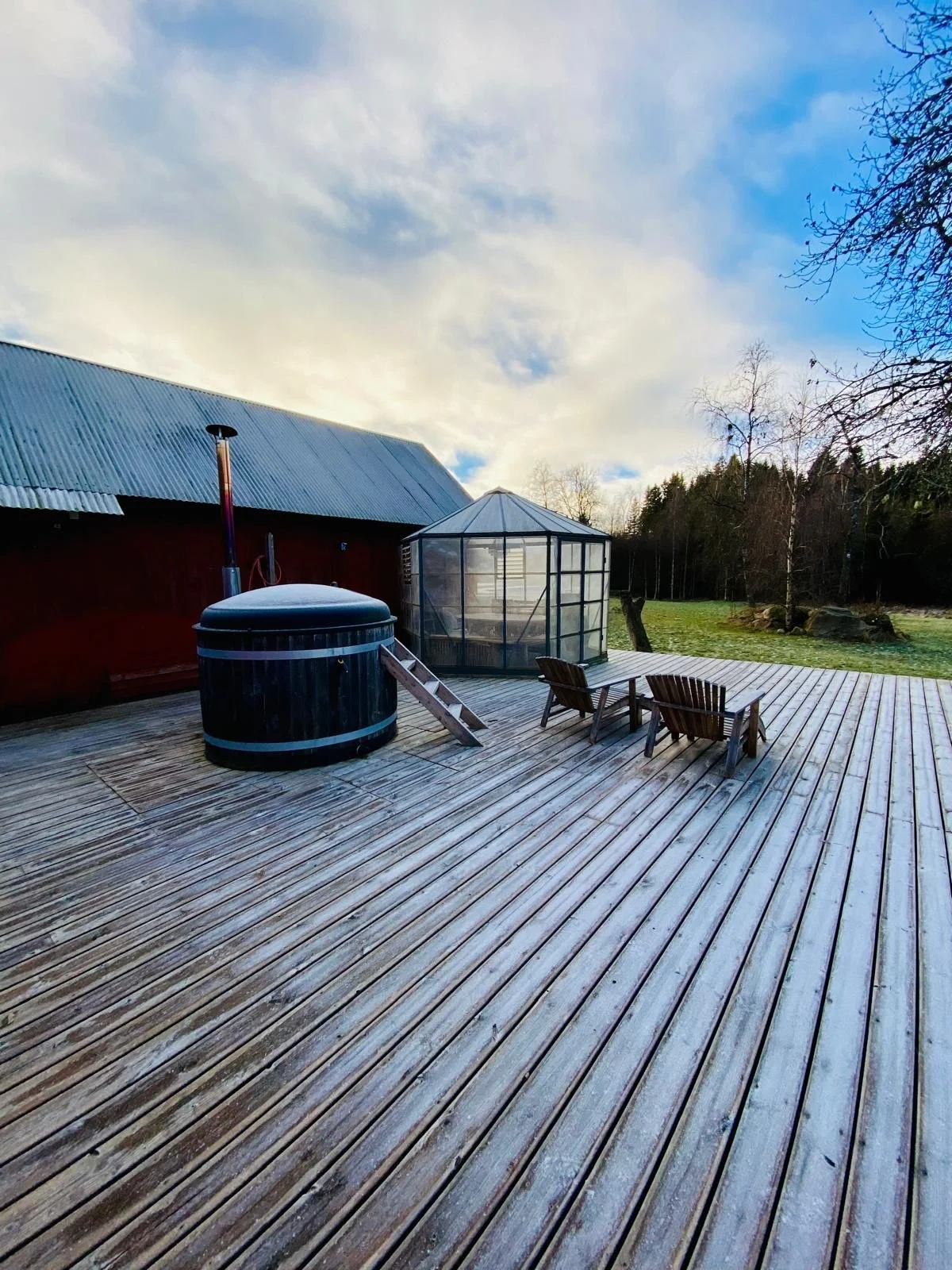 Holzterrasse mit zwei Holzsesseln, einem kleinen Whirlpool, einem Gewächshaus und einem roten Haus im Freien bei bewölktem Himmel.