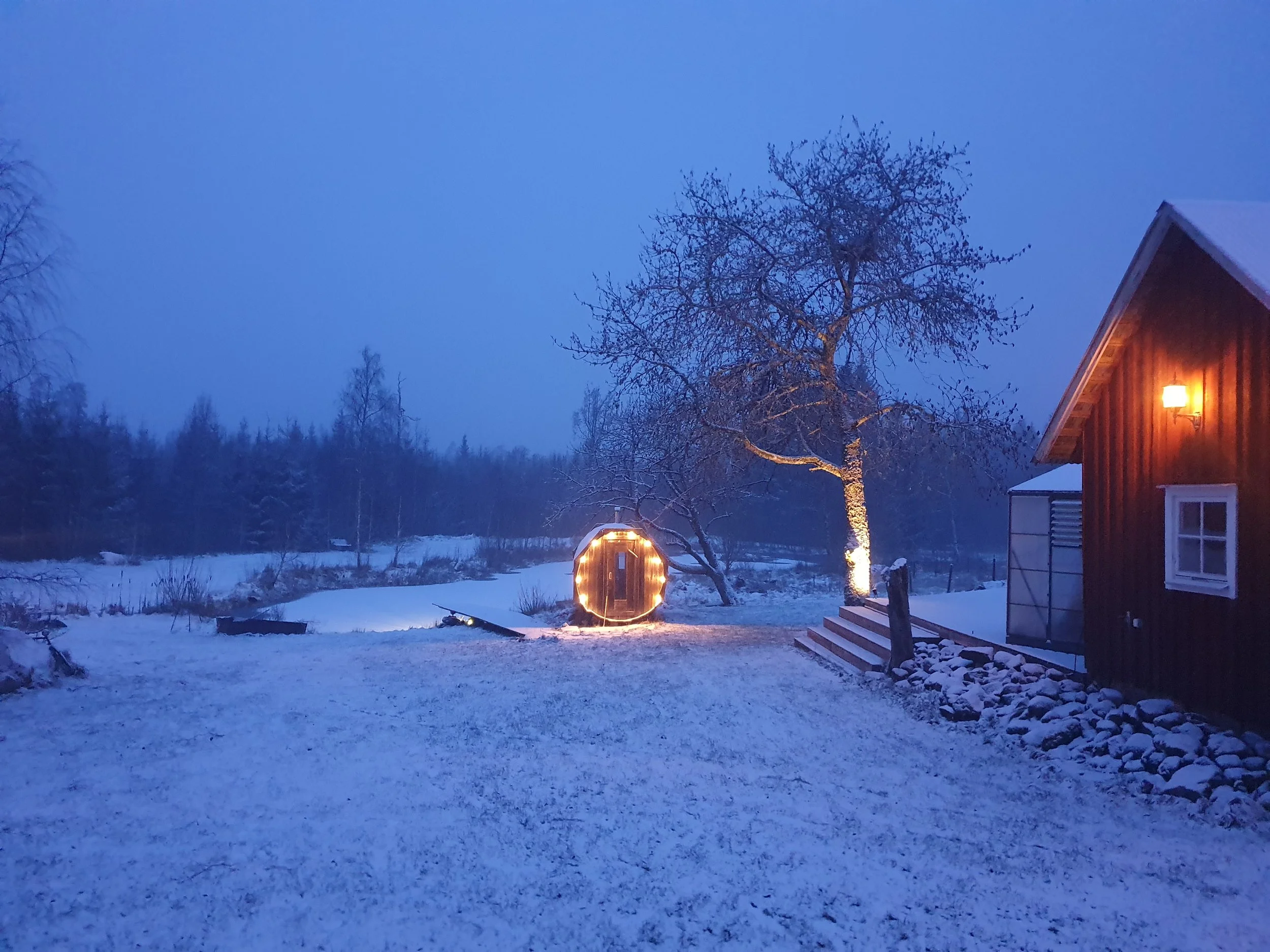 Ein verschneiter Garten bei Dämmerung mit einem Baum, einem runden Holzhaus mit Lichterkette, einem eingeschneiten Haus mit warmem Licht und einer Treppe.