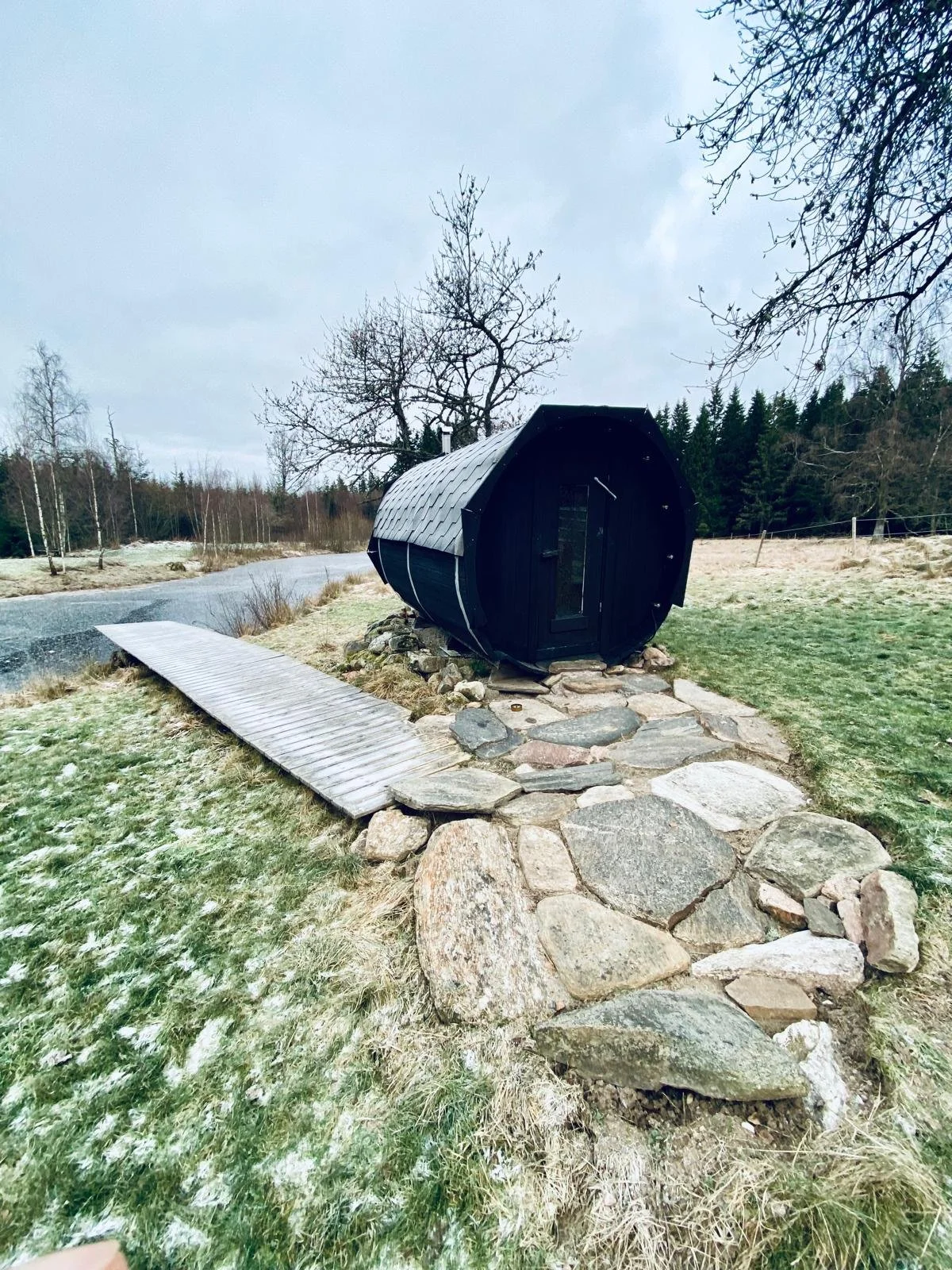 Eine kleine schwarze Sauna im Freien, umgeben von Steinen und einem Weg aus Holzplatten, in einer winterlichen Landschaft mit Bäumen und leicht gefrorenem Gras.