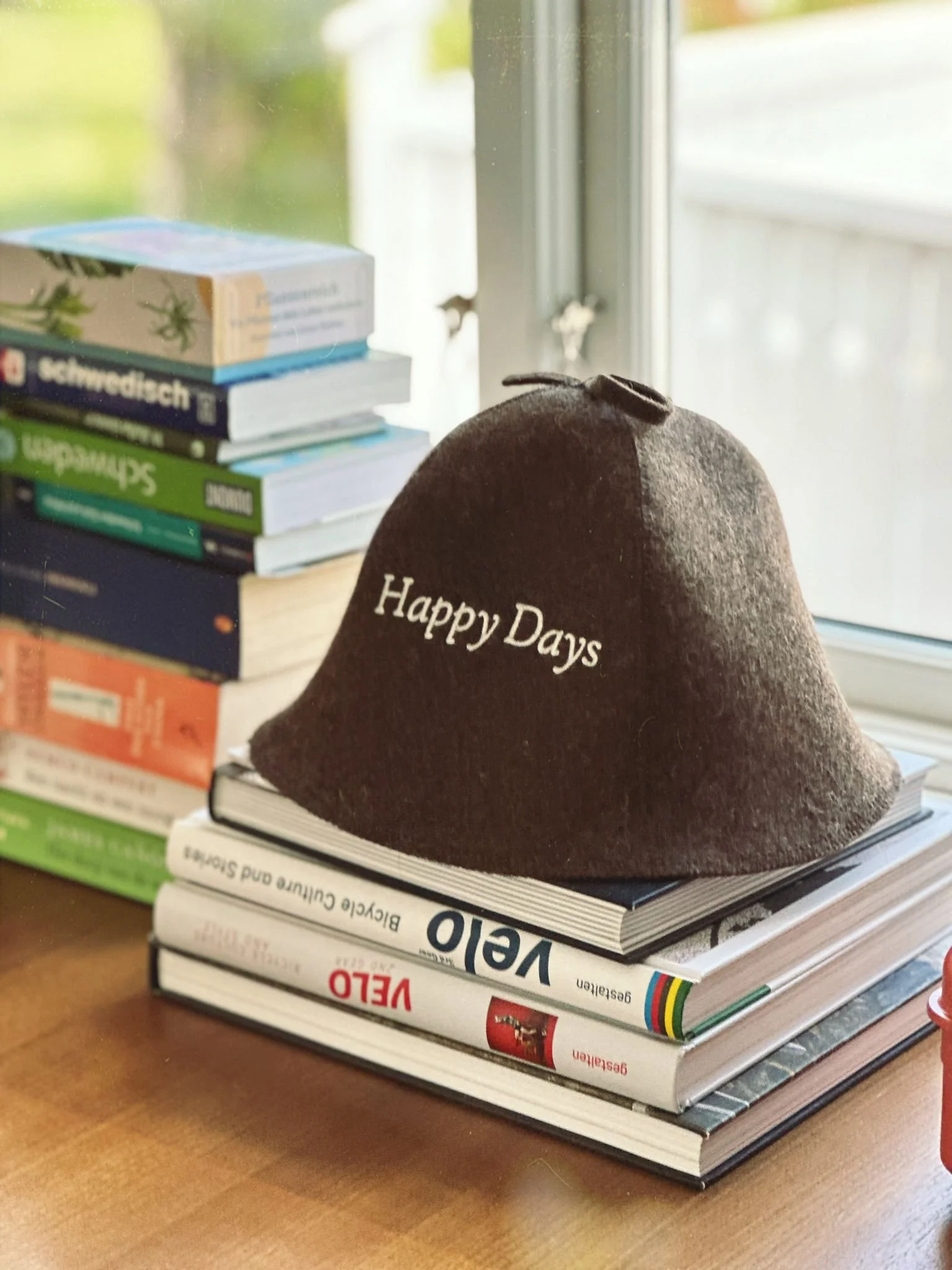 Ein brauner Filzhut mit der Aufschrift 'Happy Days' liegt auf einem Stapel von Büchern und Zeitschriften auf einem Holztisch, im Hintergrund mehrere Bücher auf einem Fensterbrett