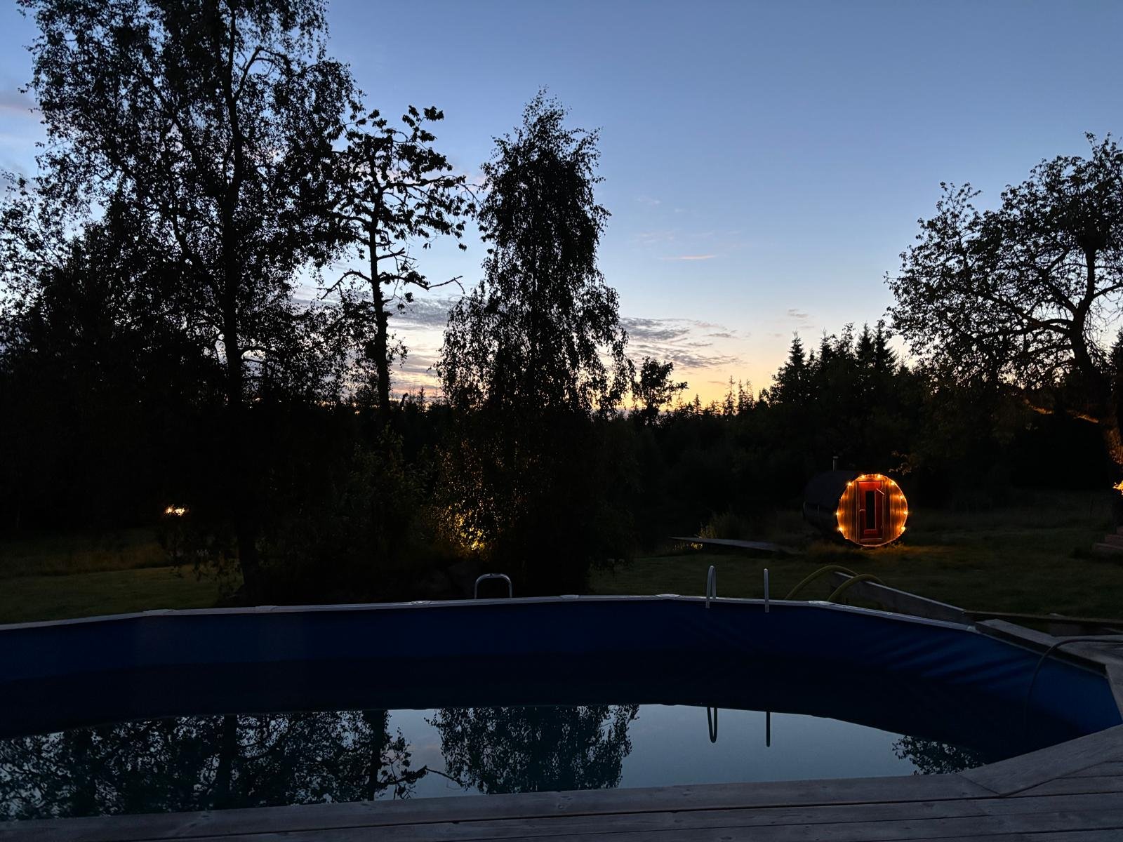 Blick auf einen Pool im Abendlicht mit Bäumen im Hintergrund und einem beleuchteten Bauwagen.