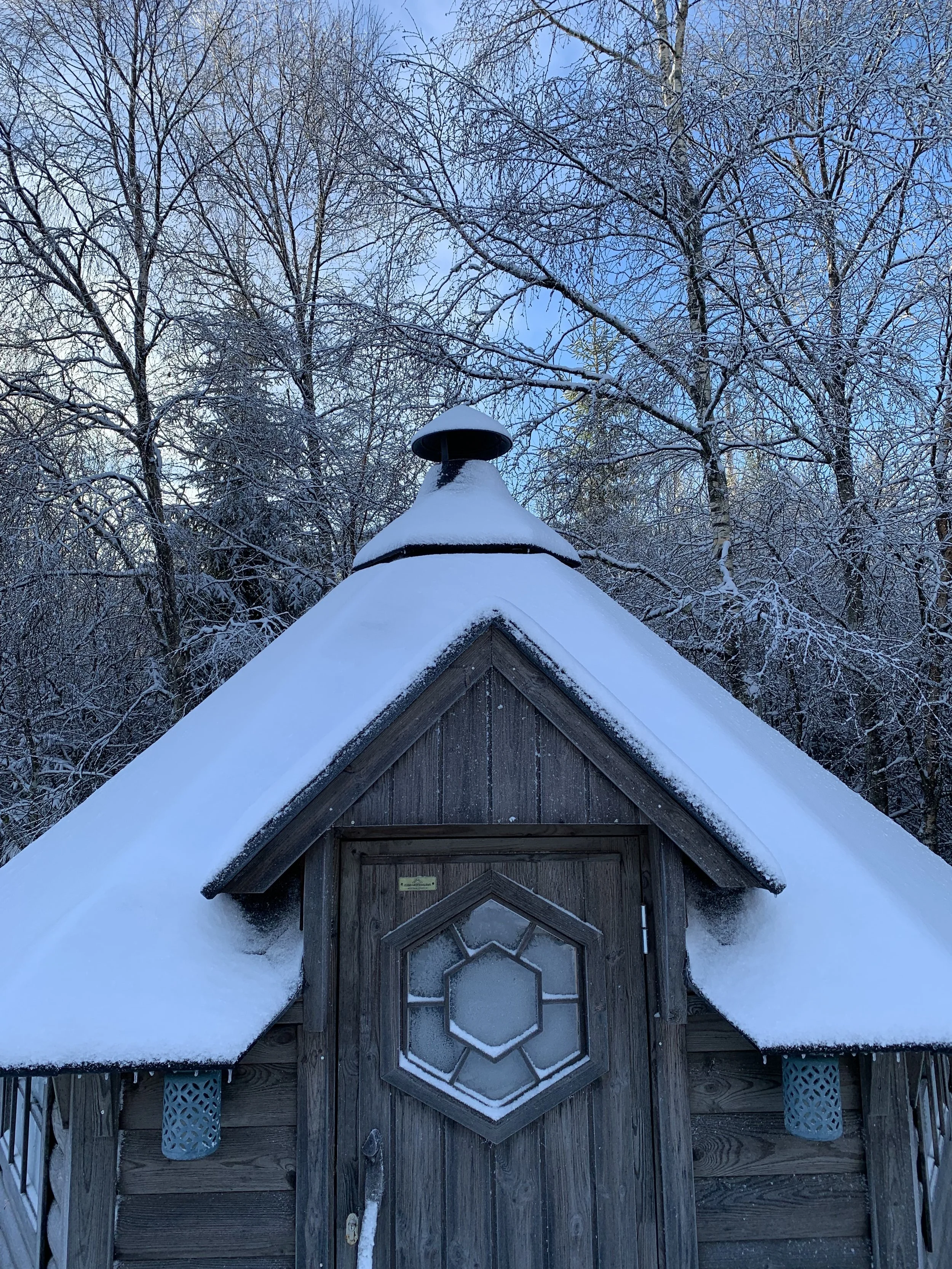 Eine kleine Holzhütte mit verschneitem Dach und einer Fliegenscheibe an der Spitze, umgeben von schneebedeckten Bäumen im Winter.