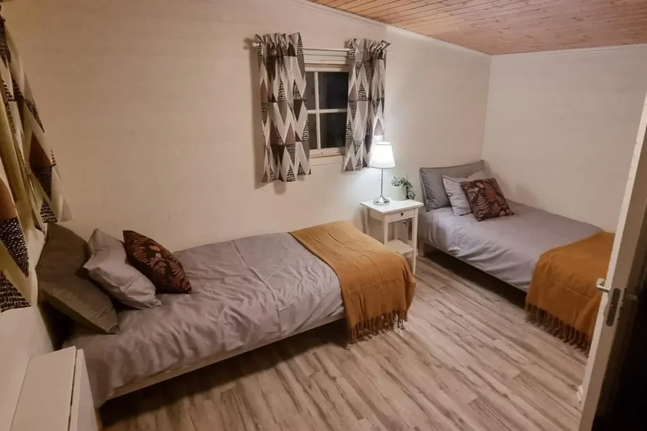 Ein Schlafzimmer mit zwei Betten, einem Nachttisch mit Lampe und Vorhängen vor einem Fenster.