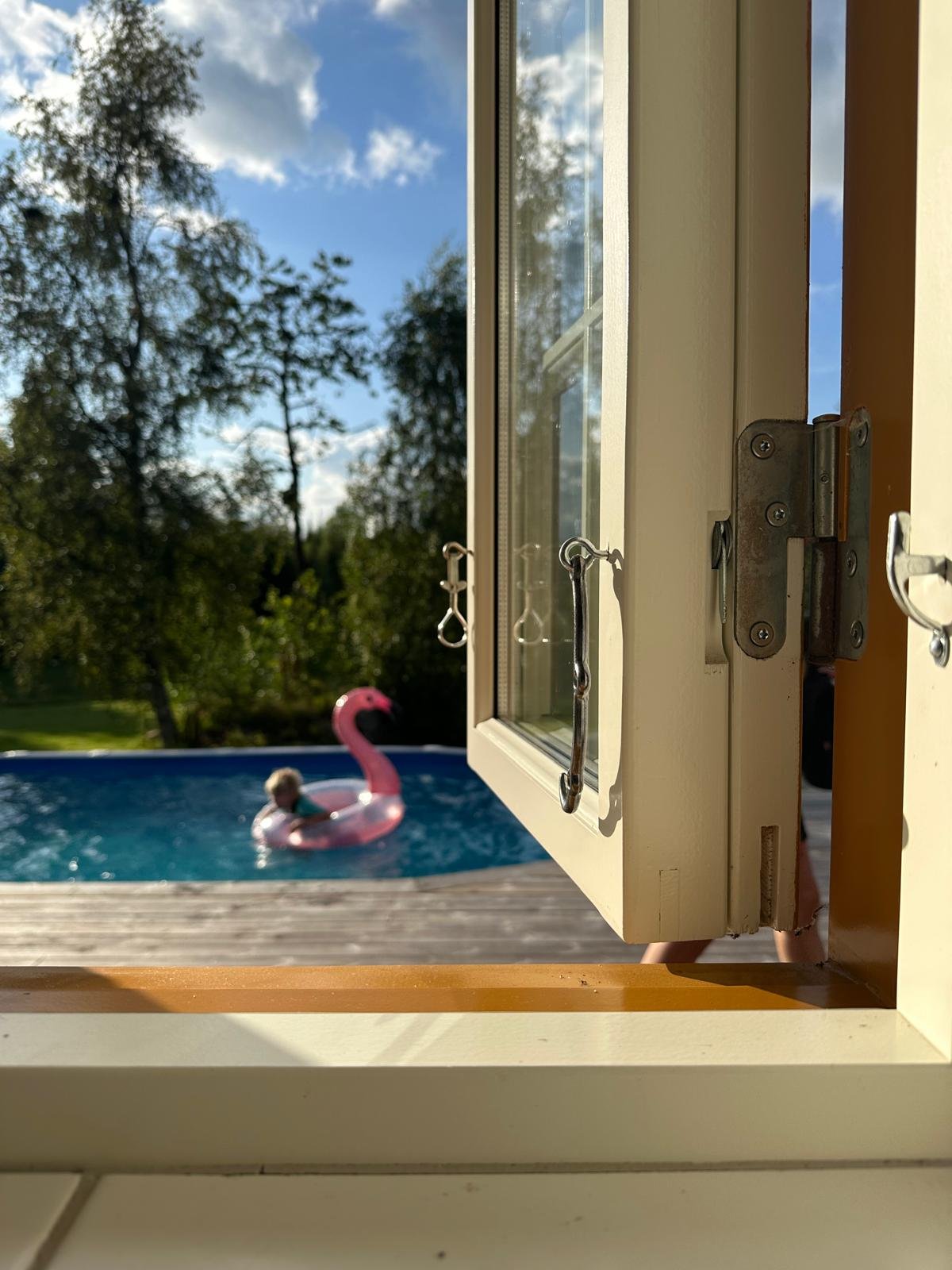 Blick durch ein geöffnetes Fenster auf einen Garten mit einem Swimmingpool und einer pinken Flamingo-aufblasung, im Hintergrund Bäume und ein blauer Himmel mit Wolken.