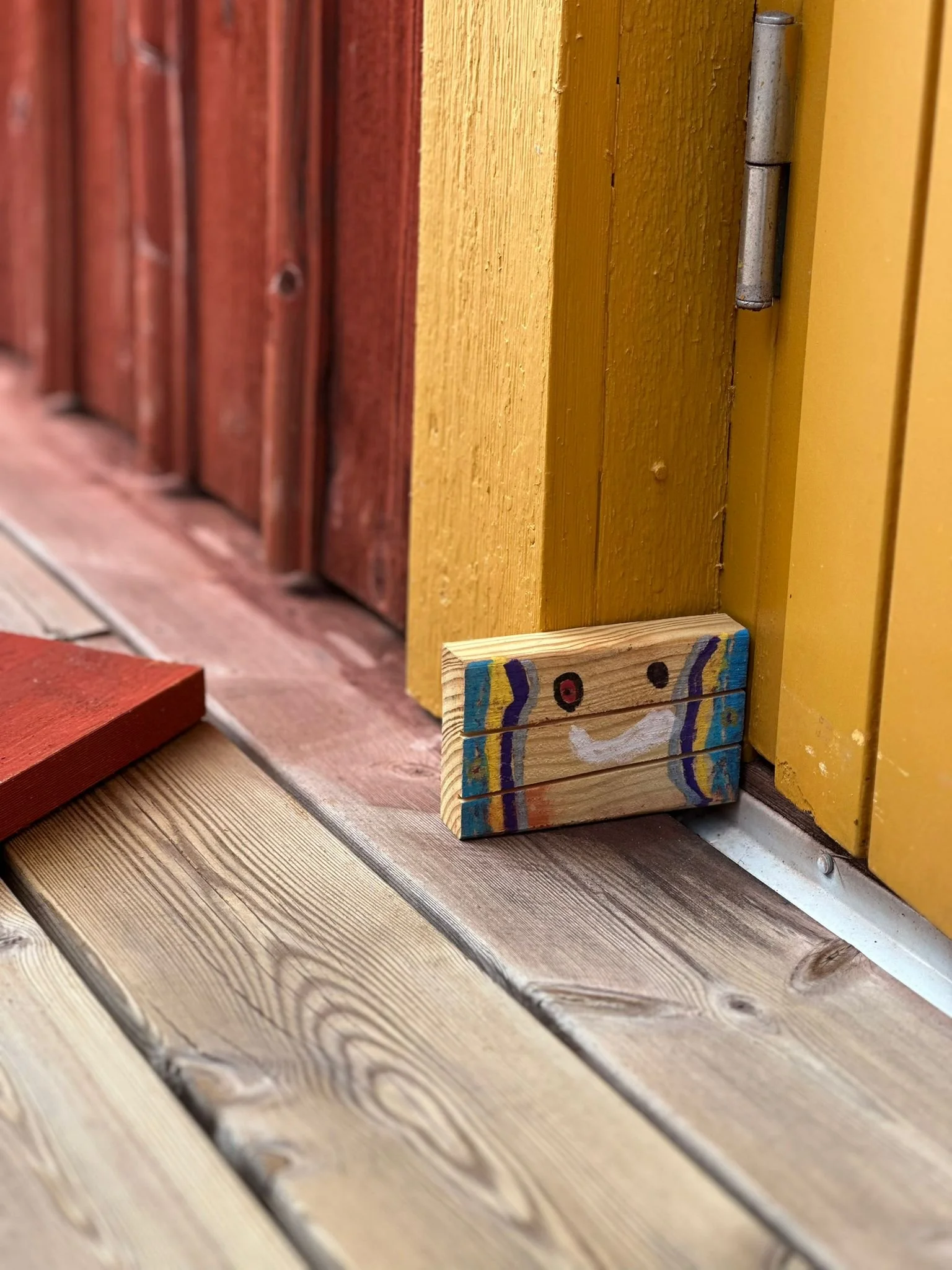 Ein kleines Holzstück mit einem lachenden Gesicht, das auf einer Holzterrasse liegt, vor einer gelben Tür und einer roten Wand.